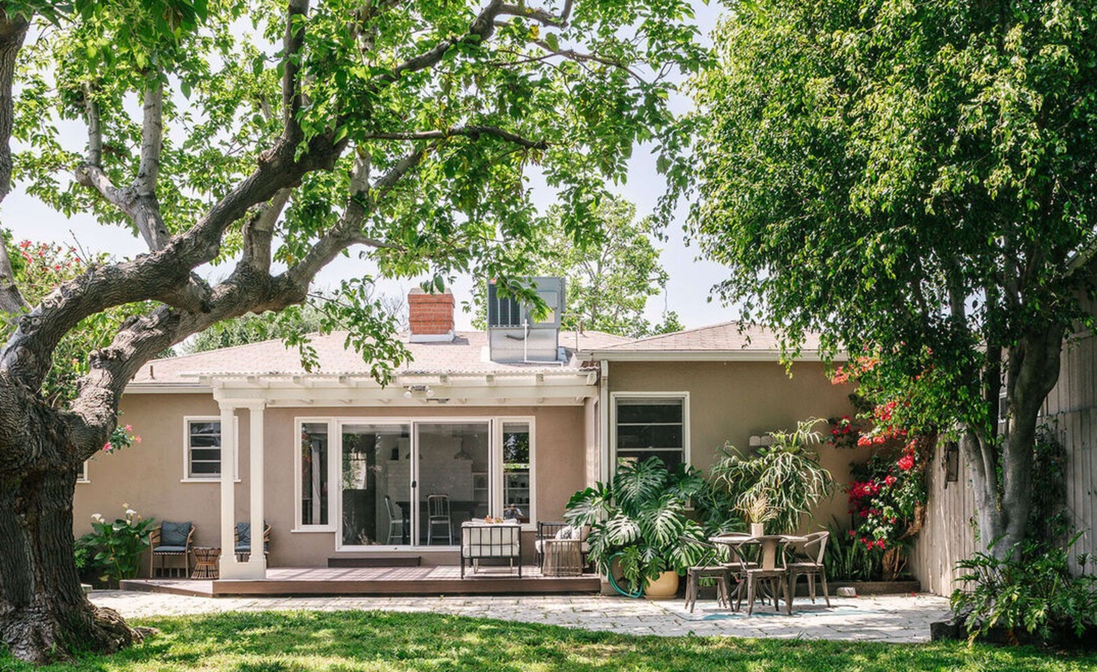 A single-story house with large windows and a deck is surrounded by trees and lush greenery in the backyard.