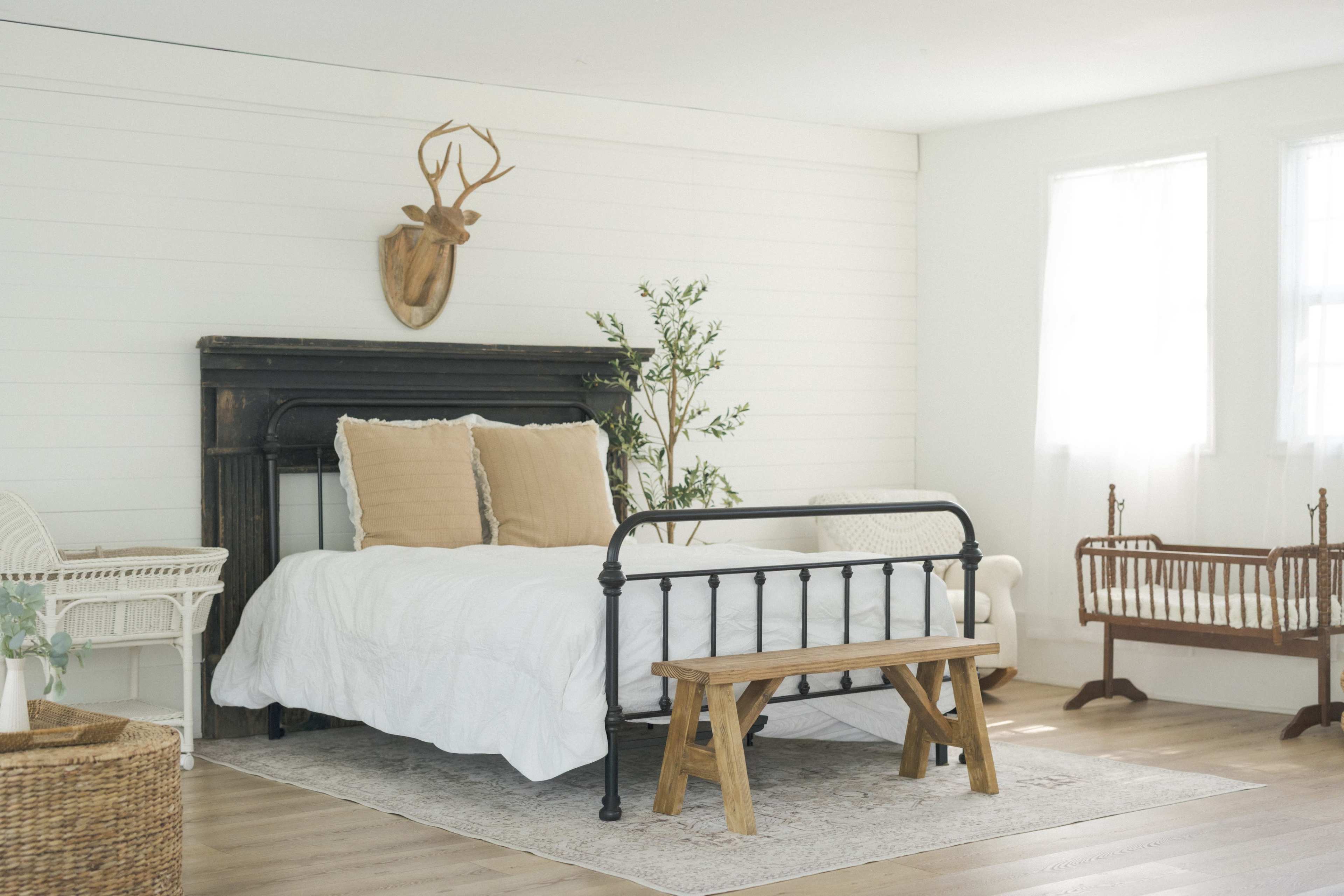 The image shows a light-filled bedroom featuring a black metal bed frame with white bedding, a wooden bench at the foot of the bed, and a small wooden cradle beside it, all set against a backdrop of white shiplap walls.