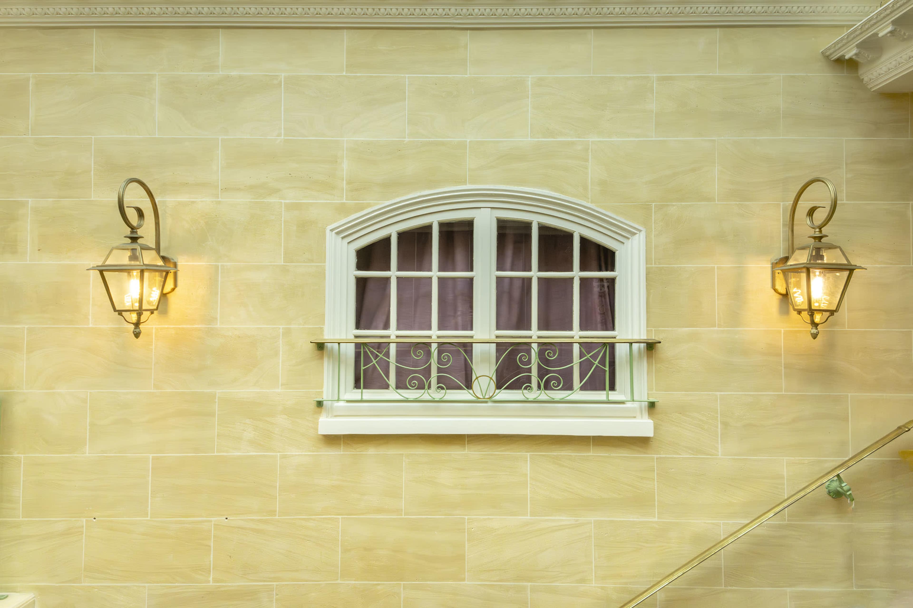 A decorative arched window framed by two wall-mounted lanterns, set against a light-colored stone wall.