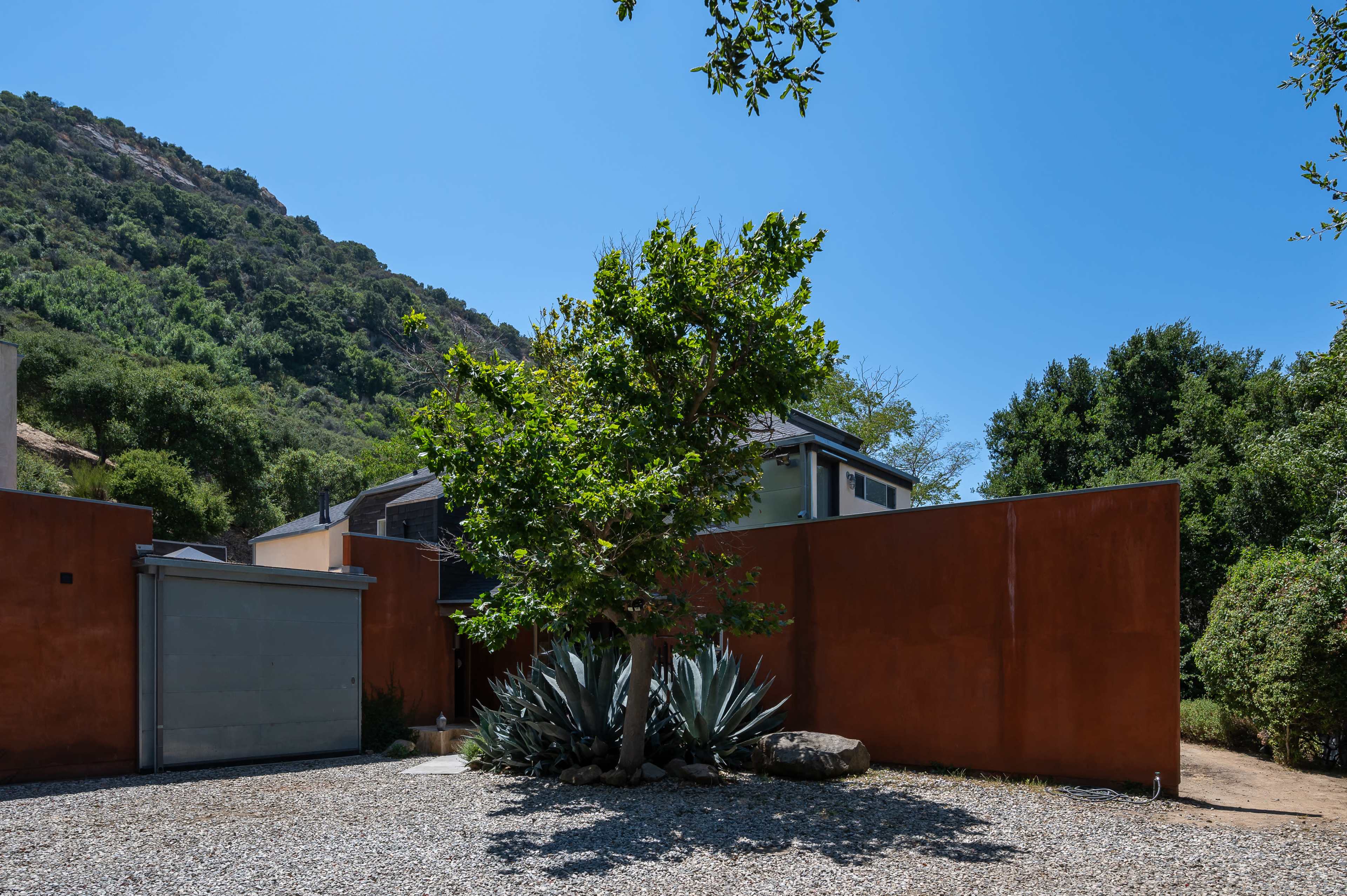 A lone tree stands next to an orange wall in a gravel landscape, with a backdrop of green hills and a clear blue sky.