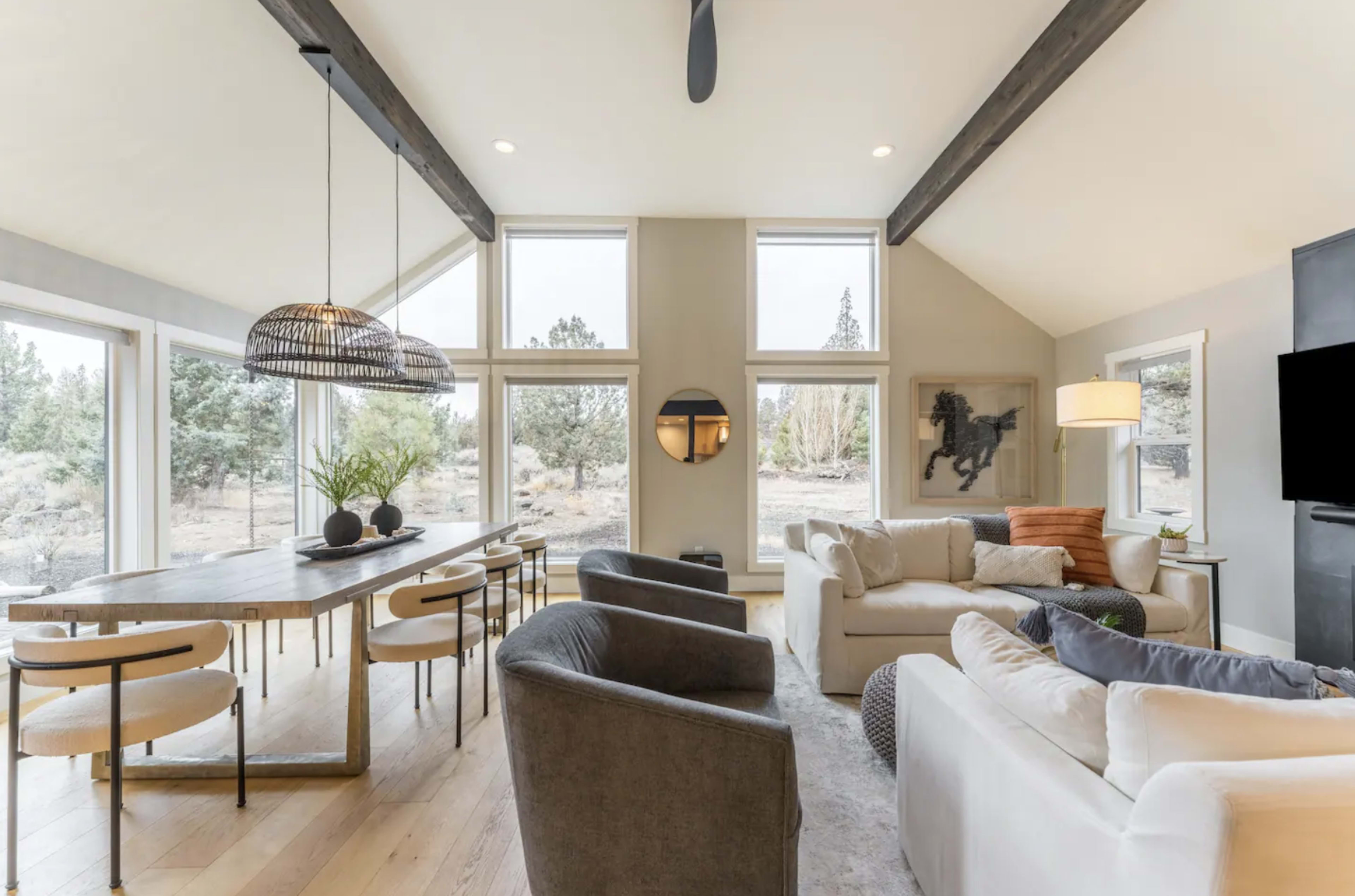 A living room features a large dining table, comfortable seating arrangements, and large windows showcasing a natural landscape.