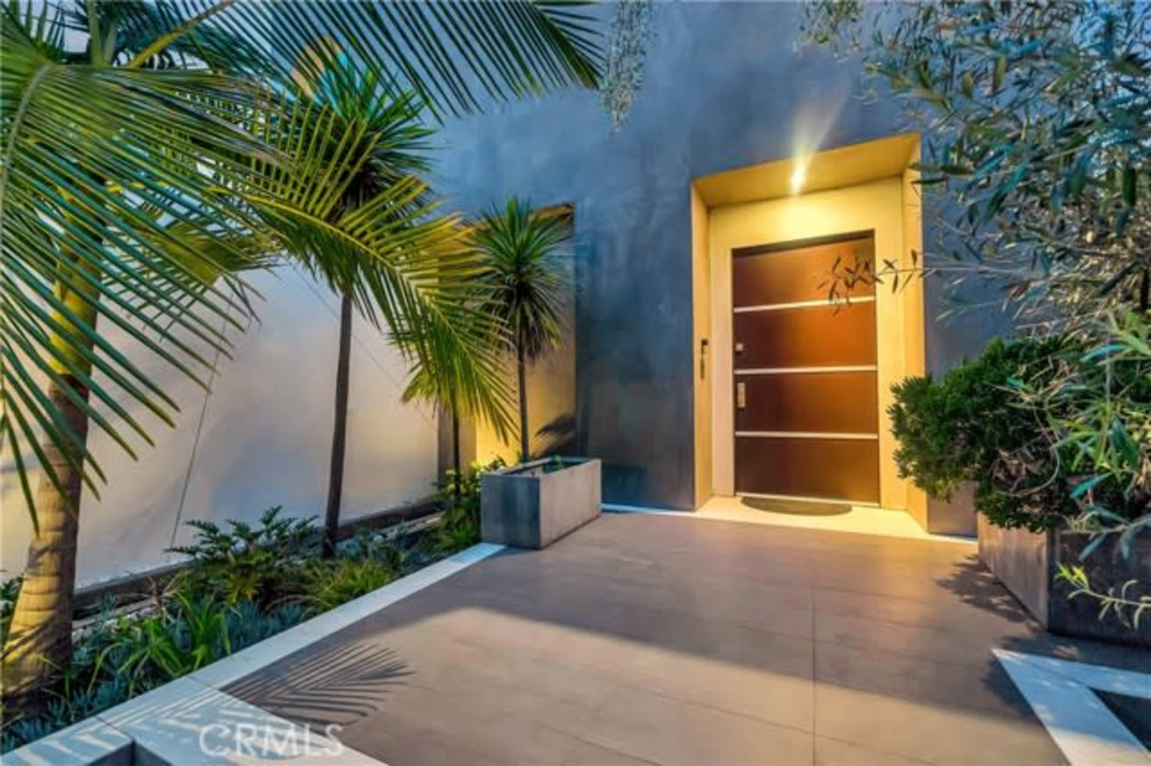 The image depicts a modern entrance with a wooden door framed by tropical plants and a well-lit pathway.