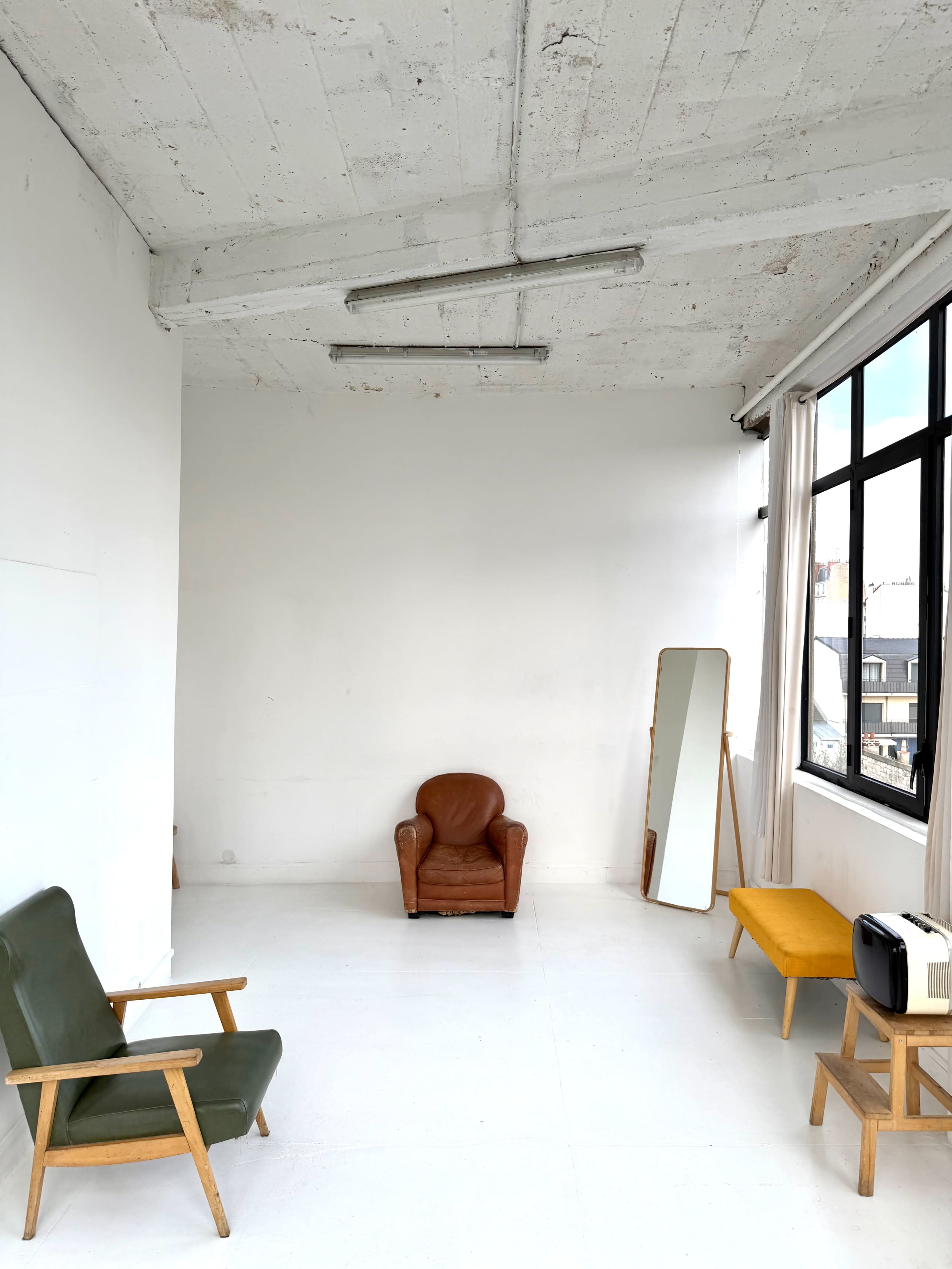 The image shows a minimalist room with a brown leather armchair, a green upholstered chair, a tall mirror, and a yellow bench near a large window.