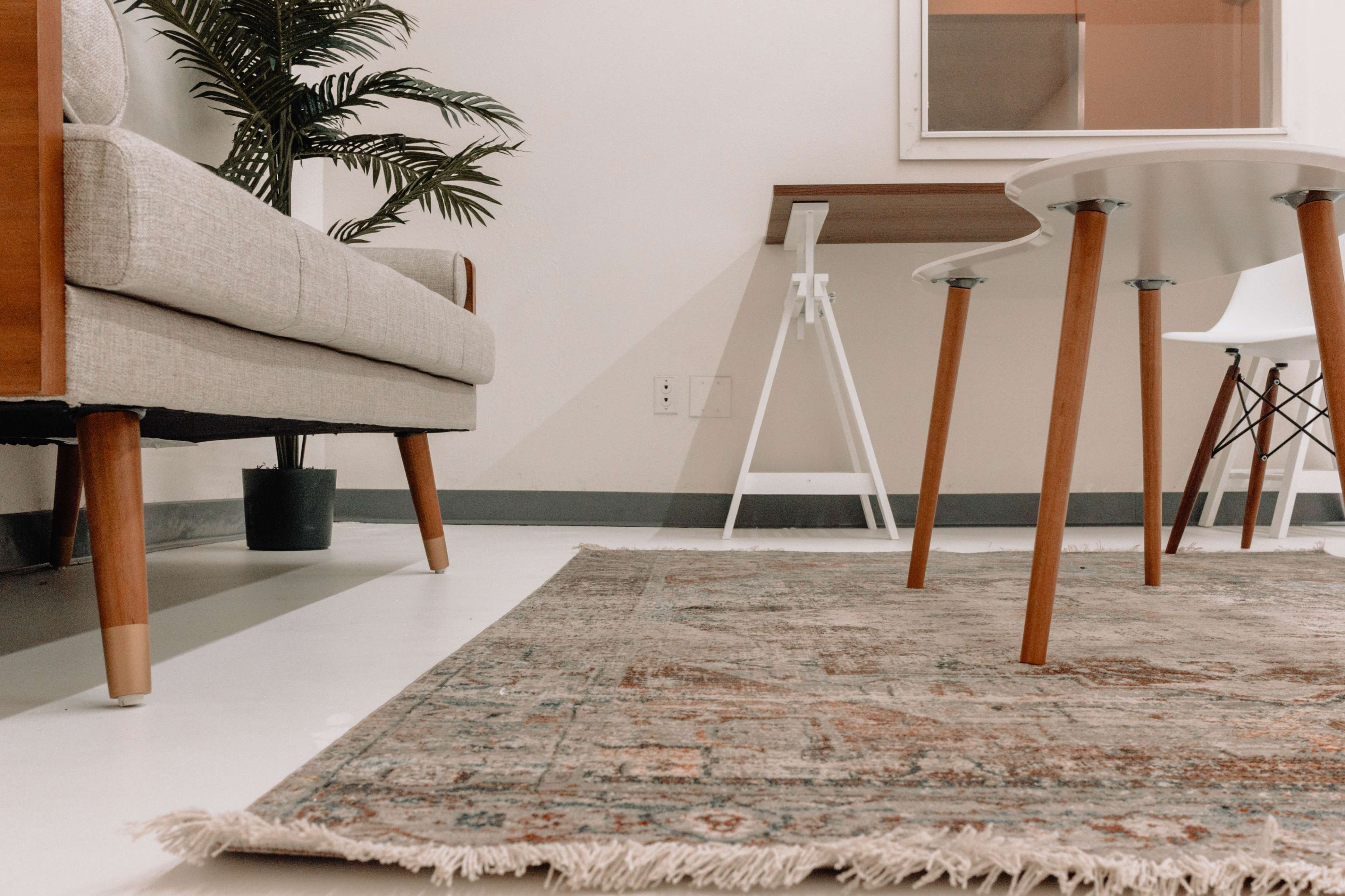 A light-colored sofa and a white table with wooden legs are positioned on a patterned rug in a minimalist room.