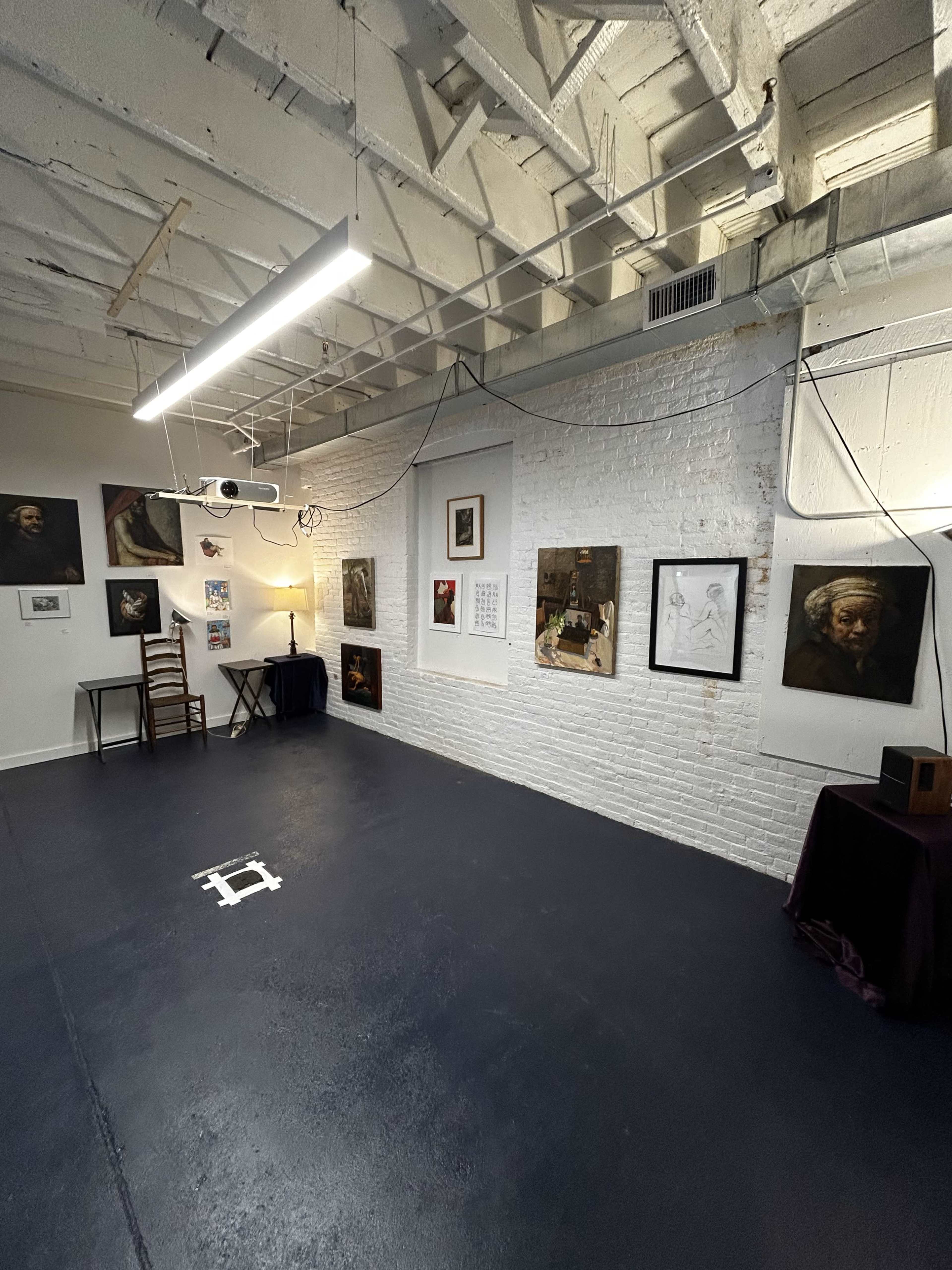 An art studio with white brick walls displaying various framed artworks, a wooden chair, and a small table set against a dark floor.