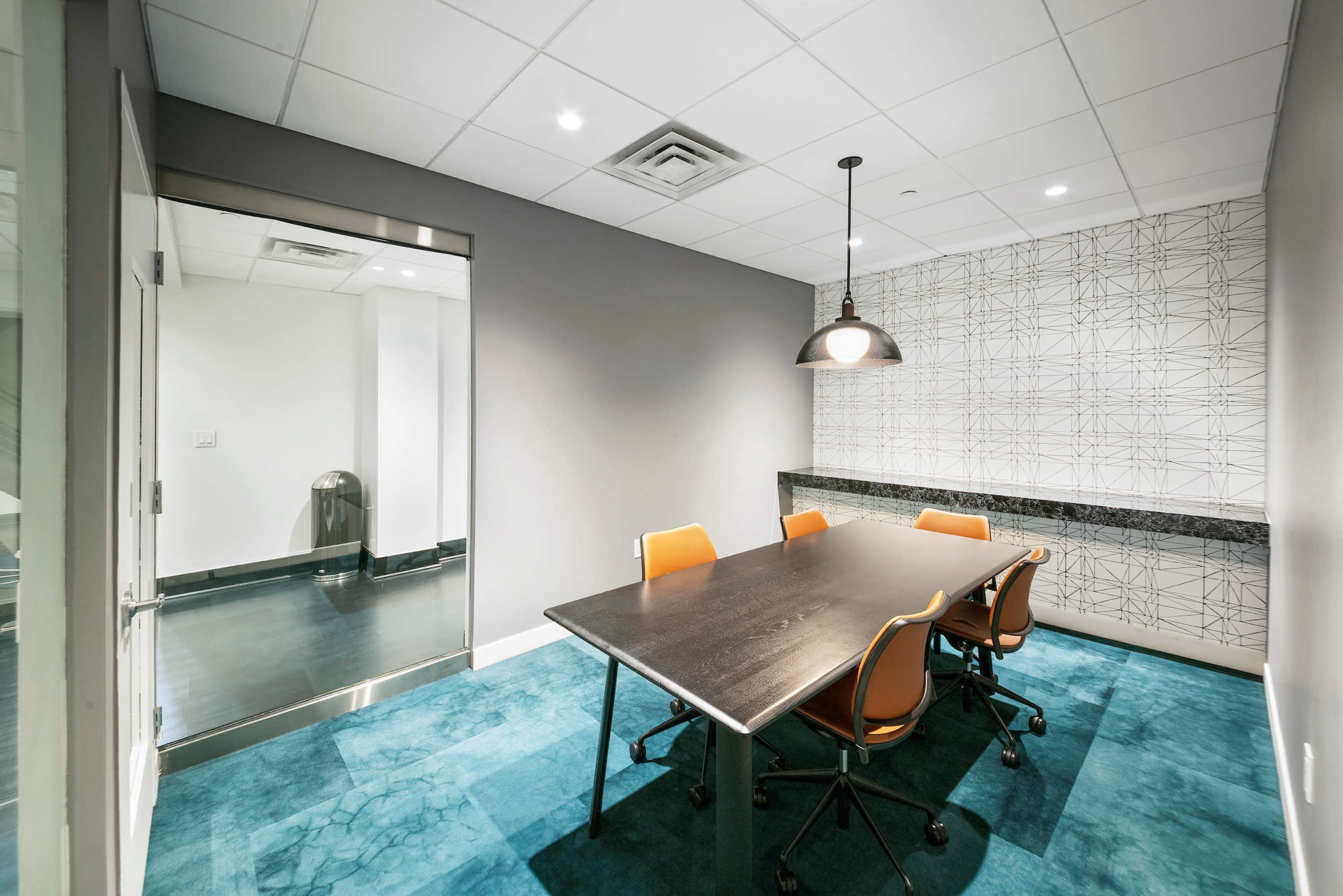 The image shows a modern conference room with a black table and orange chairs, featuring a geometric-patterned wall and a glass door leading to another area.
