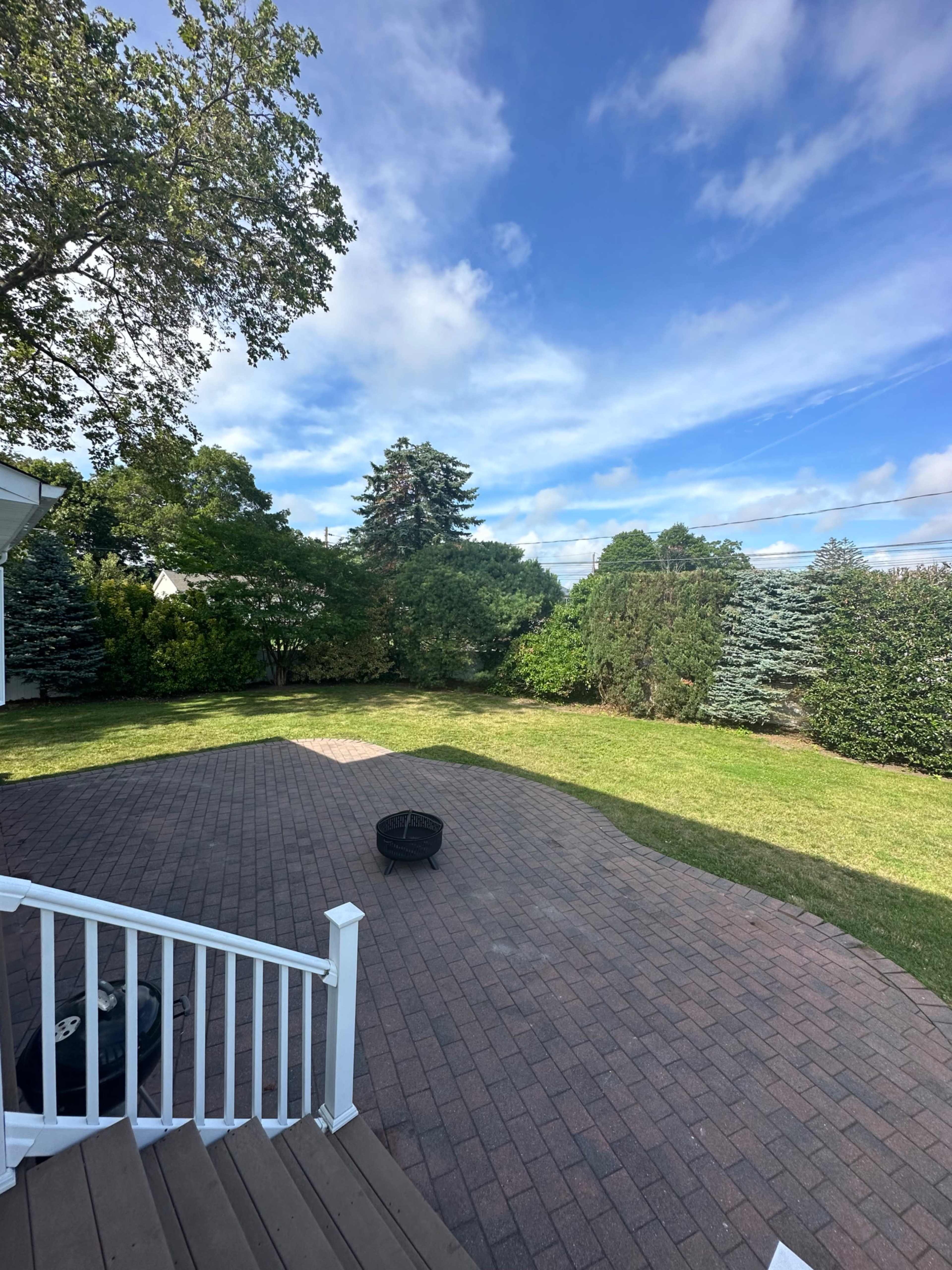 A backyard with a brick patio, green lawn, and various trees and shrubs under a partially cloudy sky.