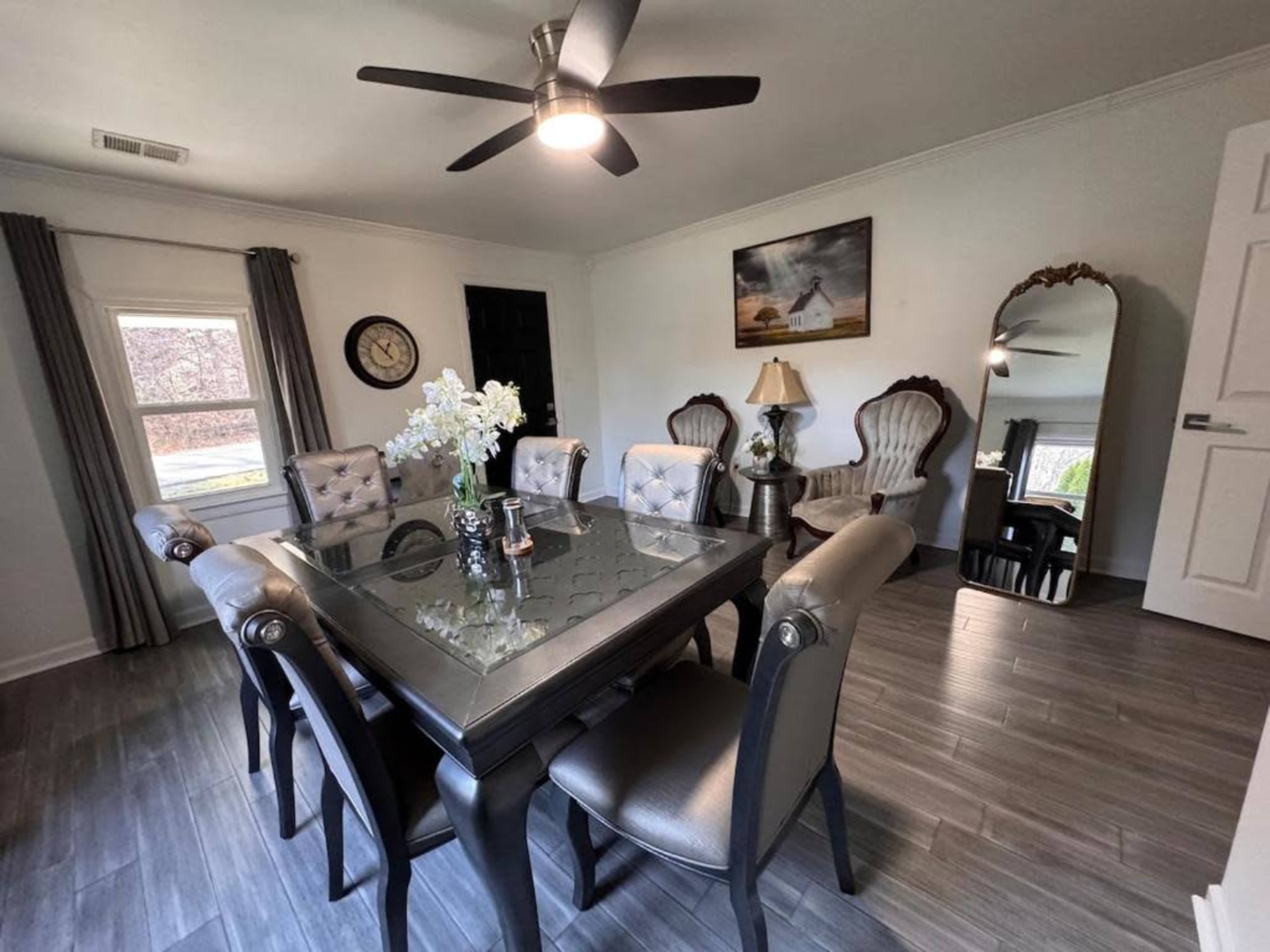A dining area features a glass-top table surrounded by upholstered chairs, with additional seating in the corner and a ceiling fan above.