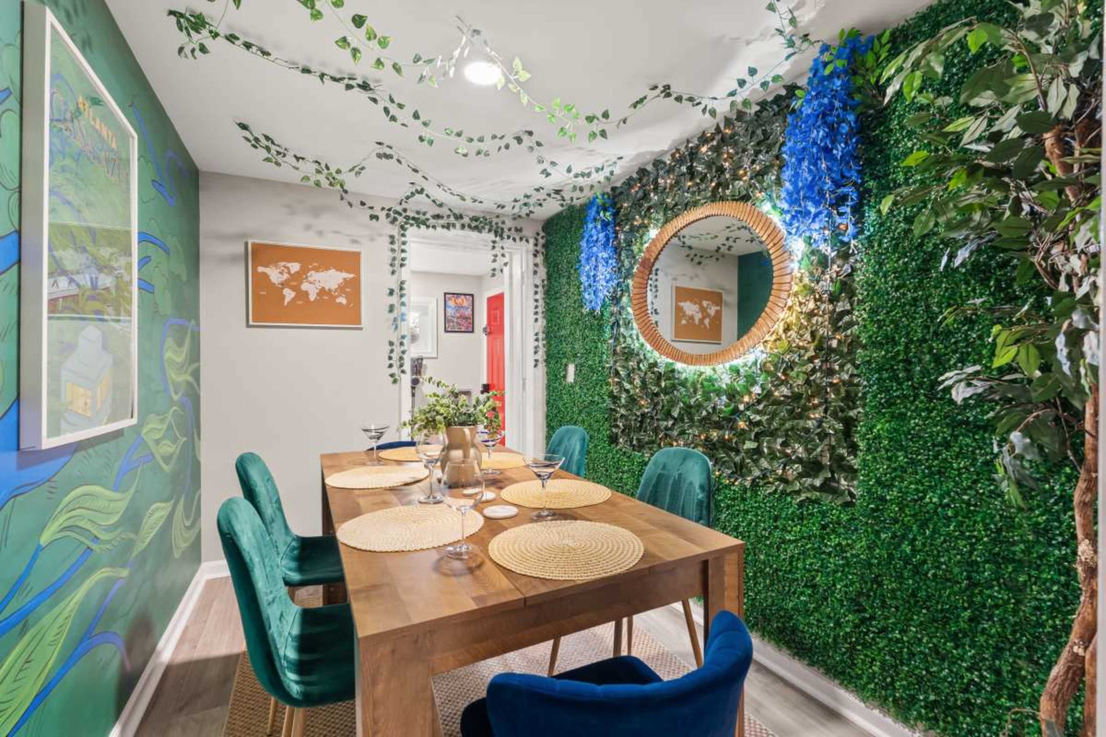 A dining area features a wooden table set with place settings, surrounded by green chairs, against a backdrop of artificial greenery and decorative art on the walls.