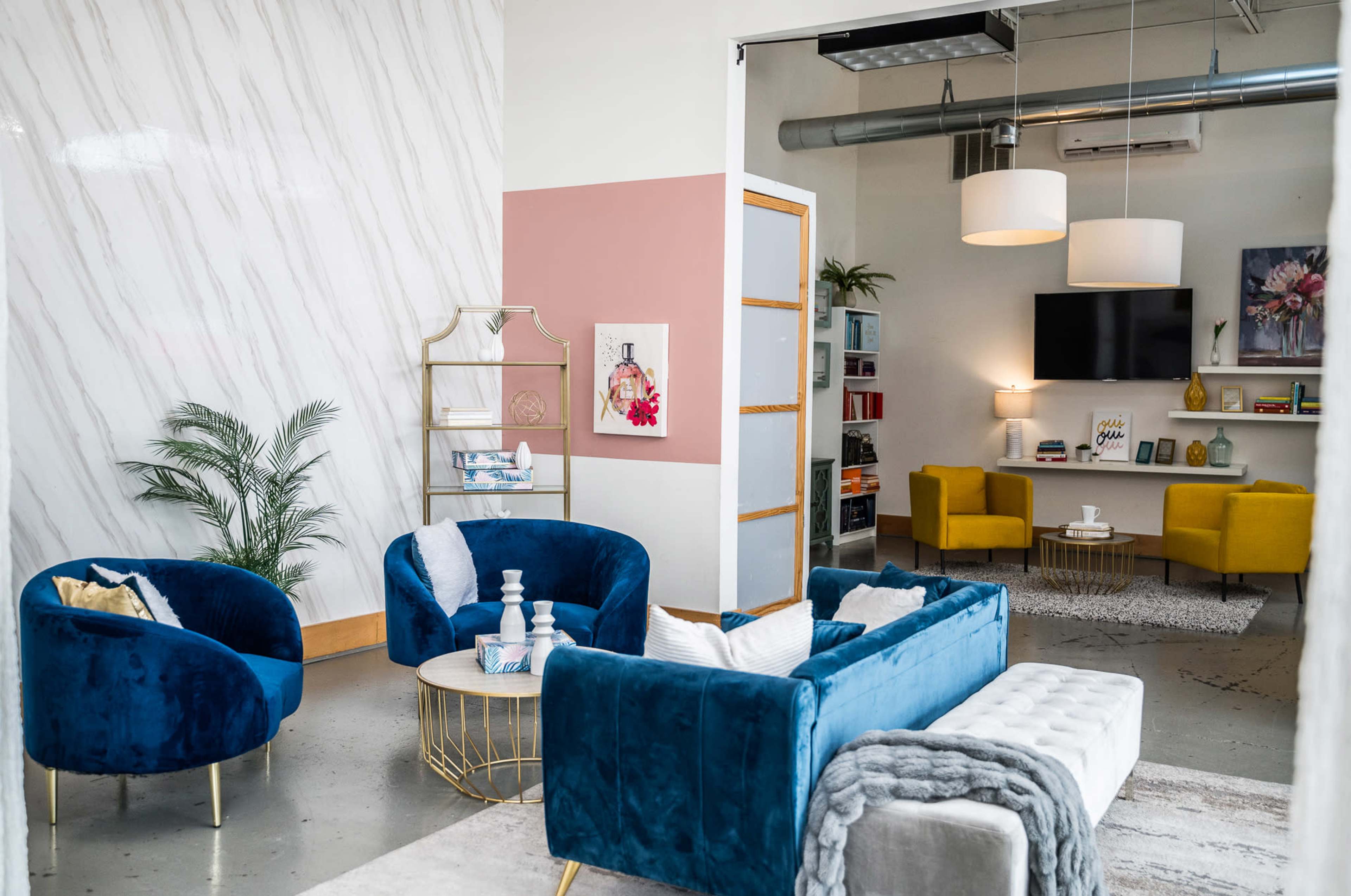 A modern living space with blue velvet chairs, a gray ottoman, a gold coffee table, and a bookshelf against a pink and white wall.