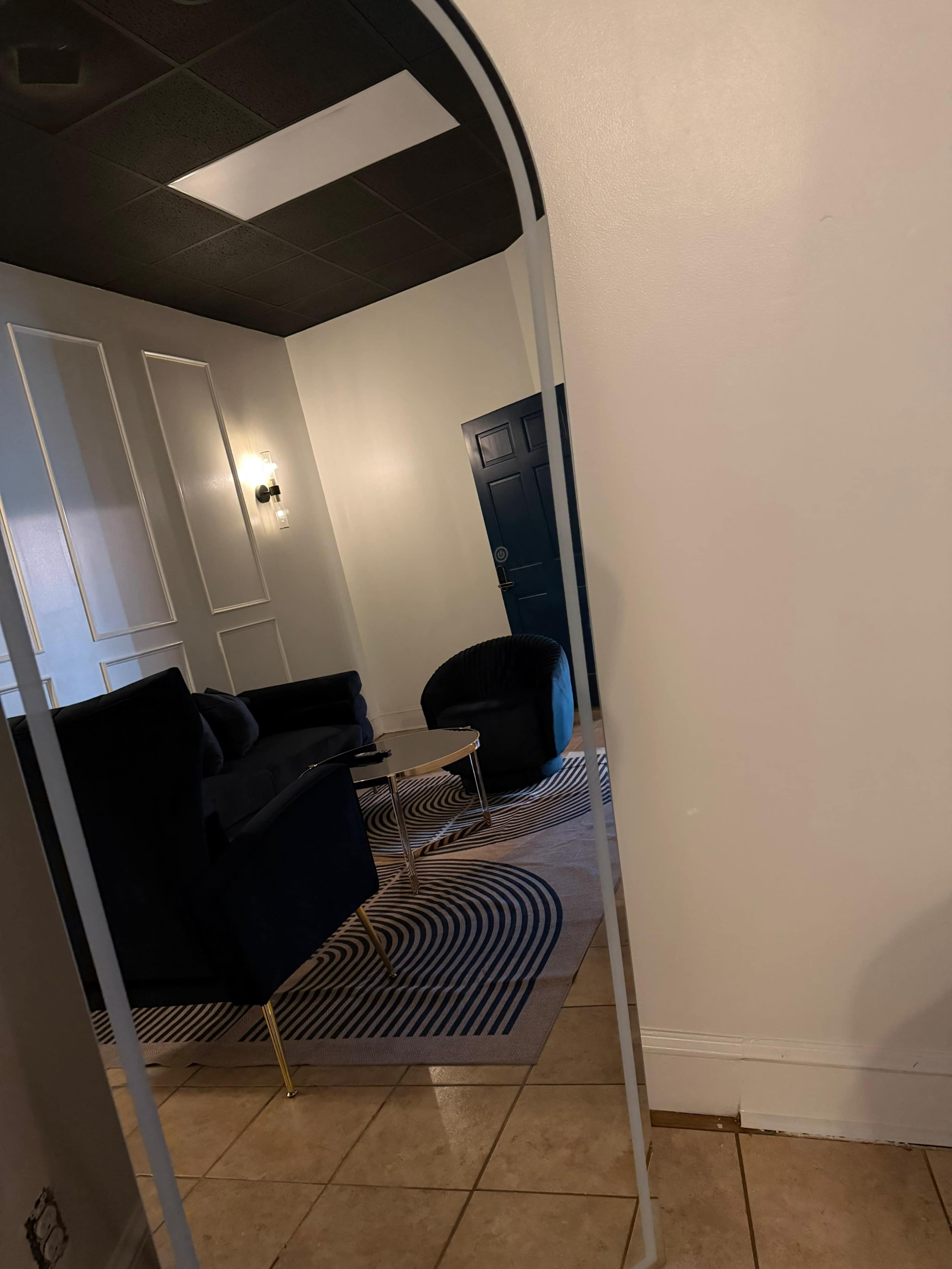 A large, arched mirror reflects a stylish room with a dark sofa, round table, and a blue door.