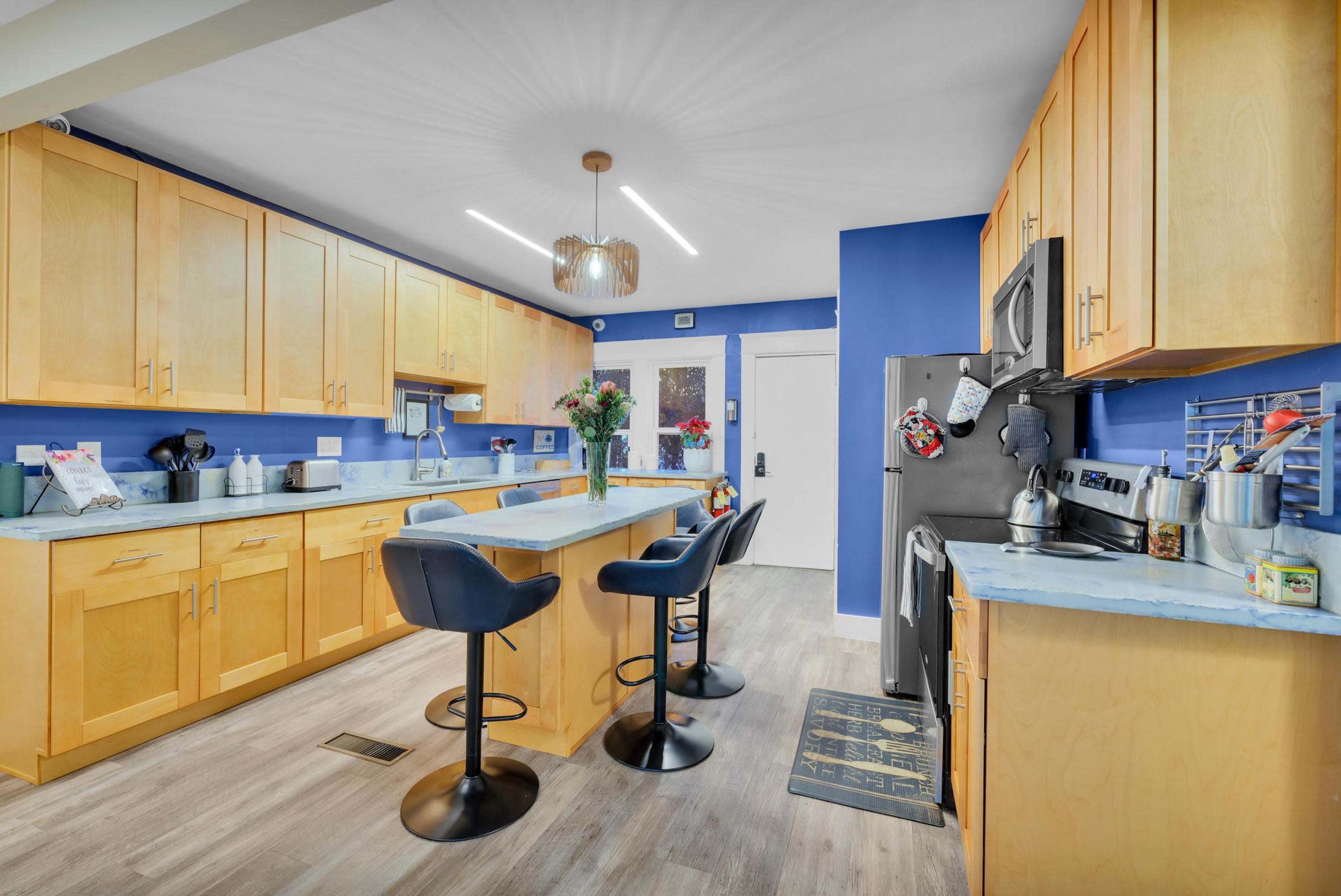 A modern kitchen with light wooden cabinets, a blue accent wall, and a central island with three black bar stools.
