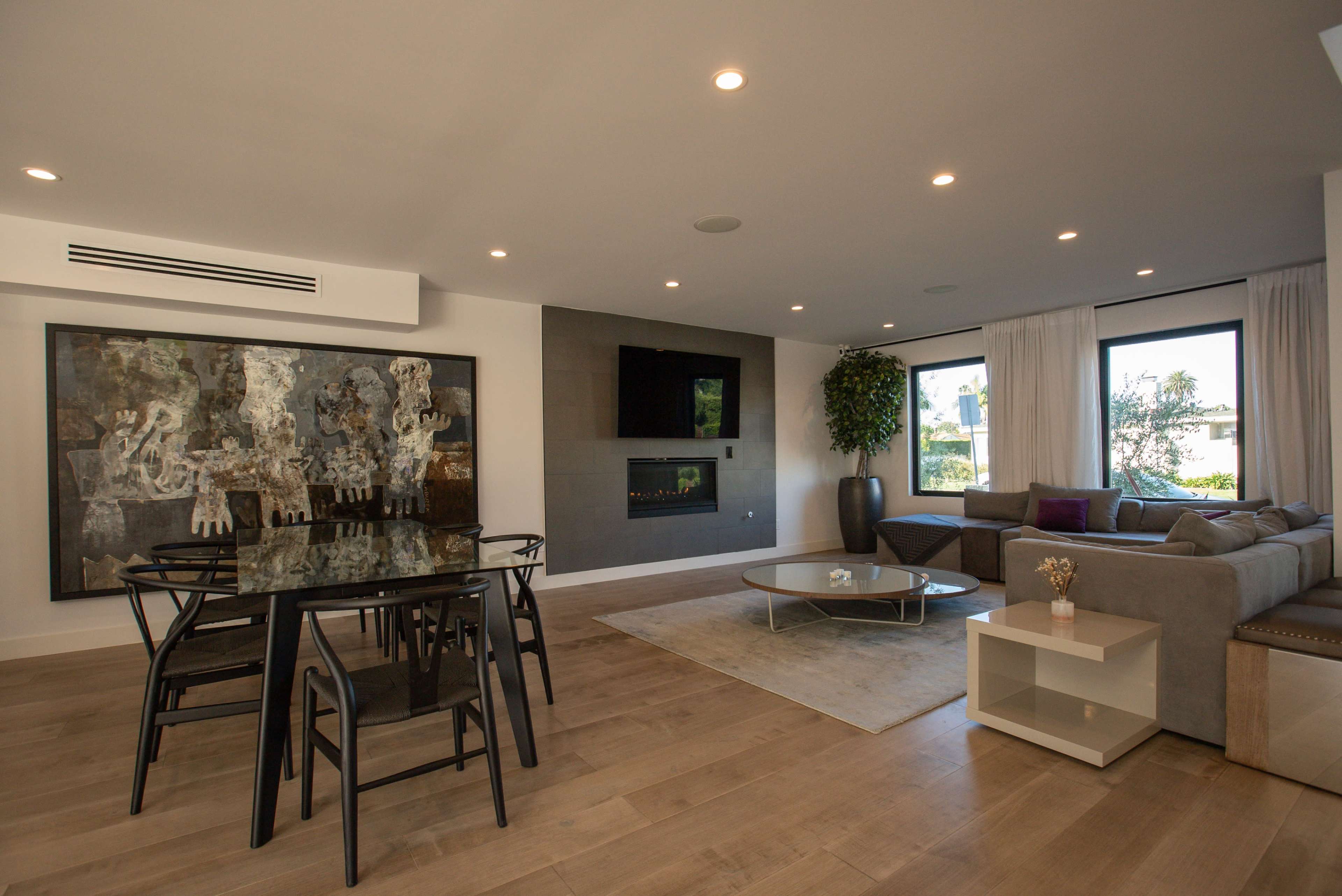 A modern living room features a dining table with black chairs, a sectional sofa, a wall-mounted fireplace, and large windows with natural light.