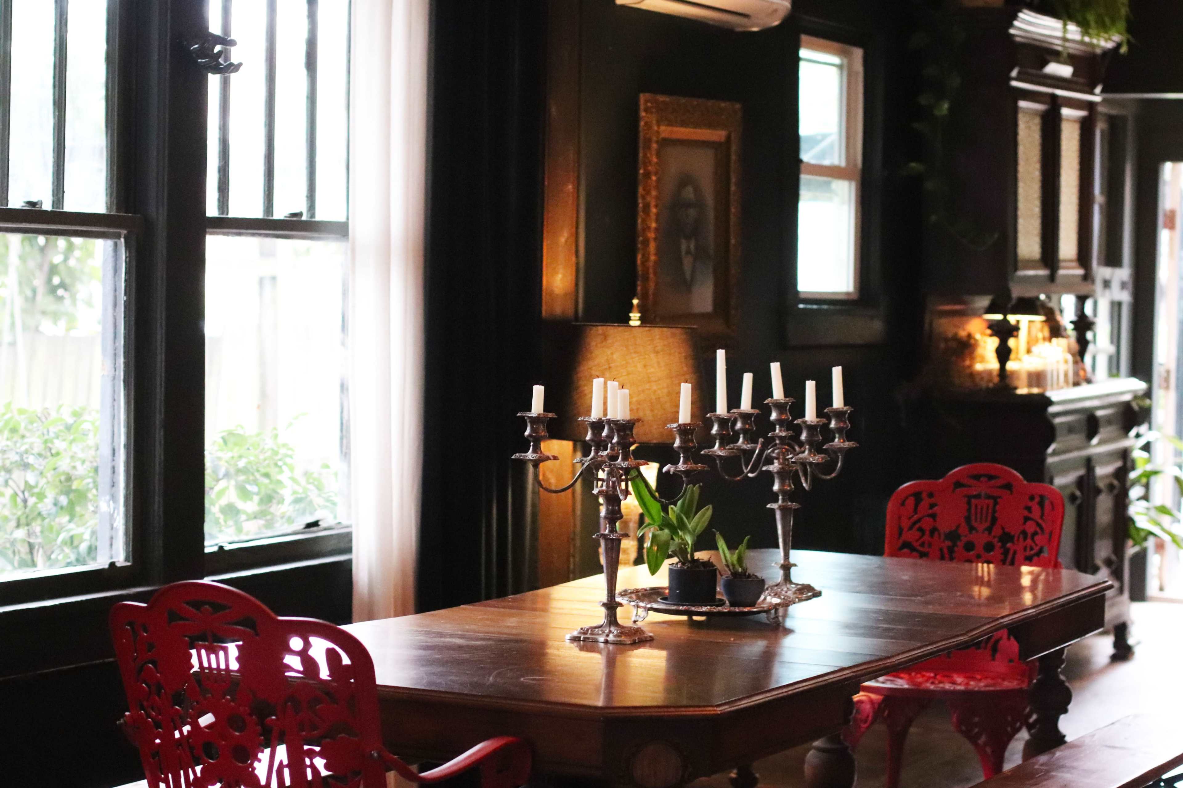 The image shows a dining area with a large wooden table, red chairs, a candelabra with candles, and a plant centerpiece, set against a dark wall and windows.