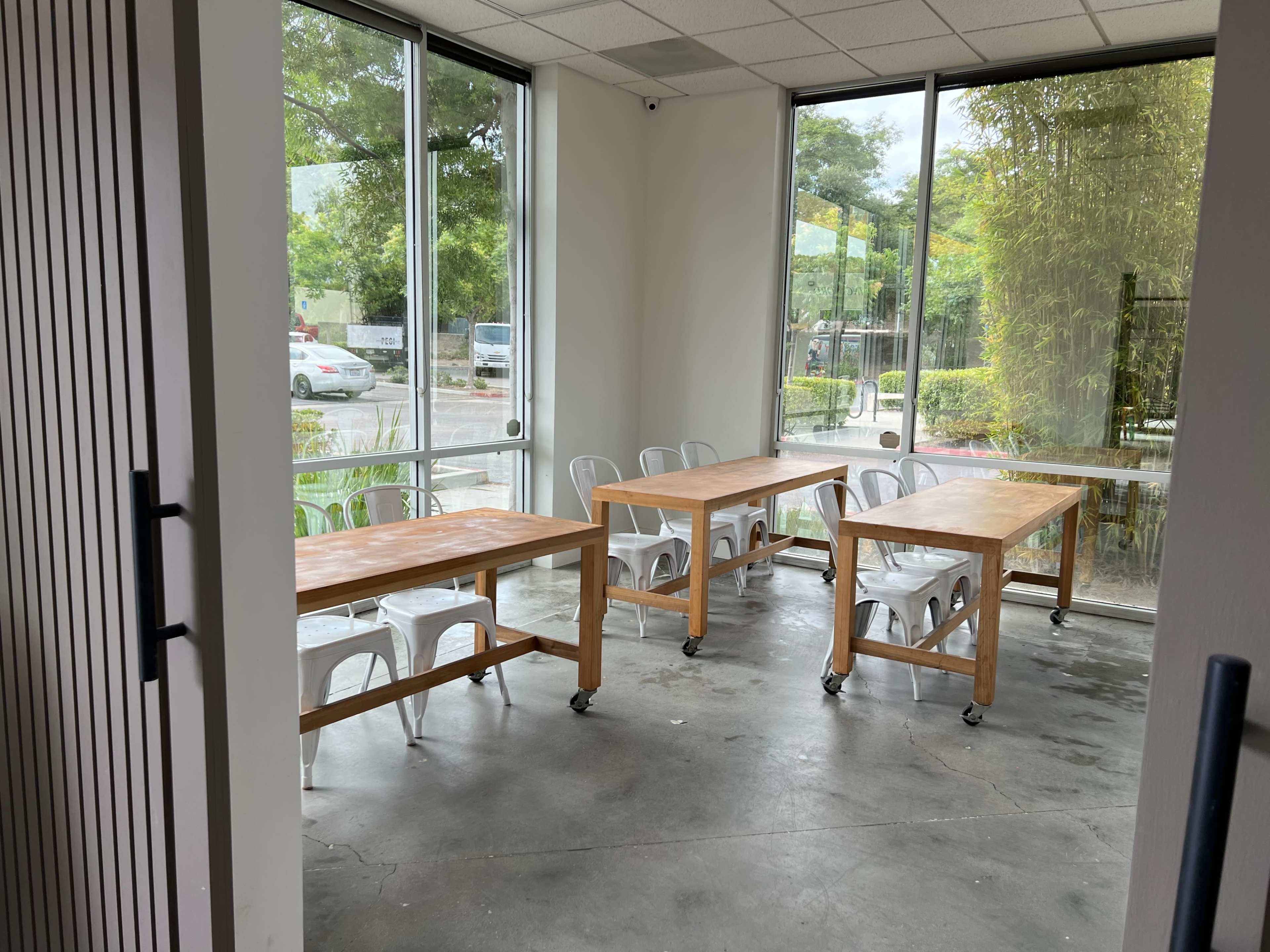A bright room with large windows, featuring four wooden tables on wheels and white chairs arranged in a simple layout.