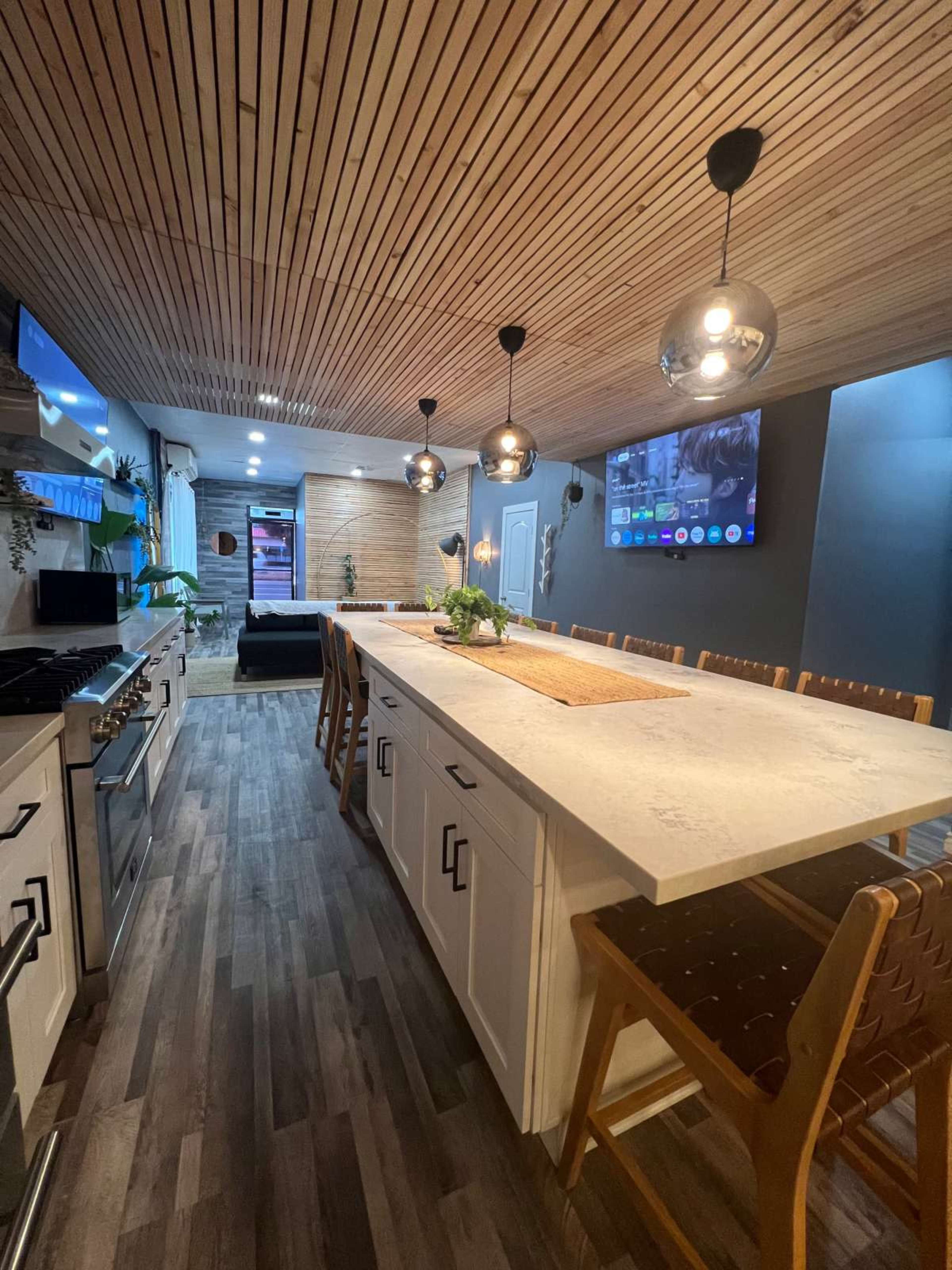 A modern kitchen with a long island, pendant lighting, wood paneling on the ceiling, and a wall-mounted television.