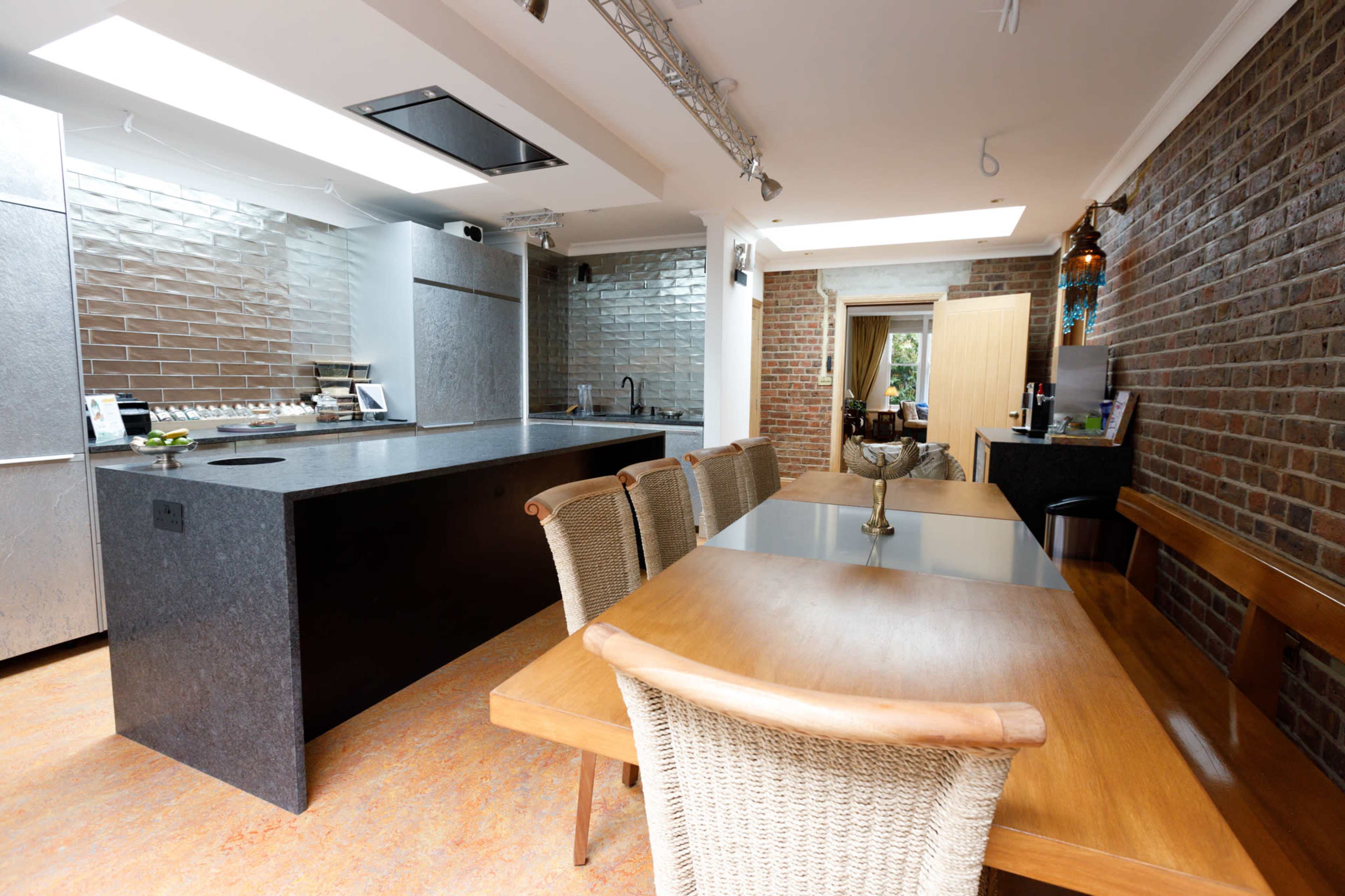 The image depicts a modern kitchen with a dark countertop, a wooden dining table, and exposed brick walls, featuring a sleek design and ample lighting.