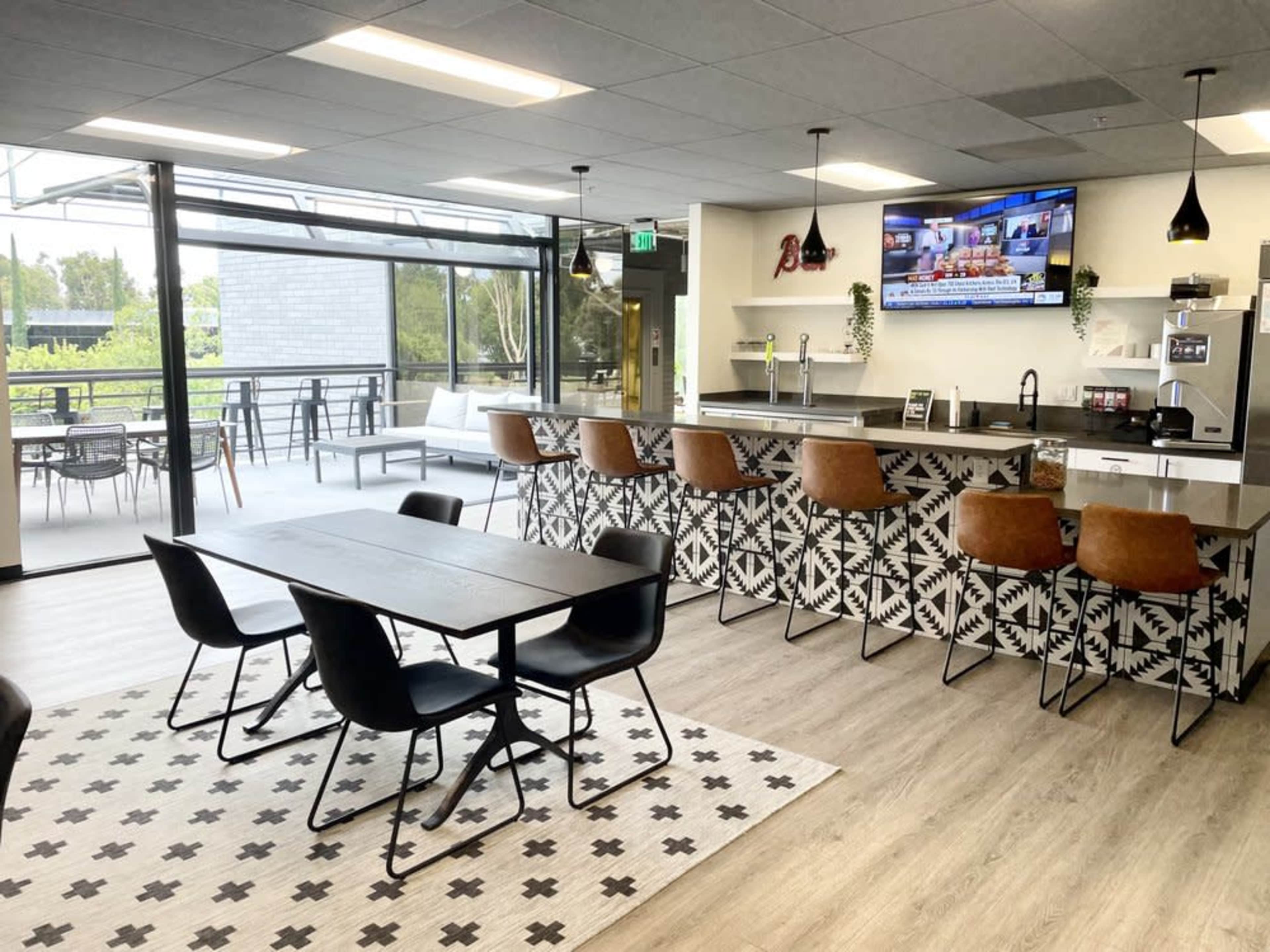 The image shows a modern kitchen and dining area with a countertop, bar seating, a dining table, and large windows overlooking an outdoor space.