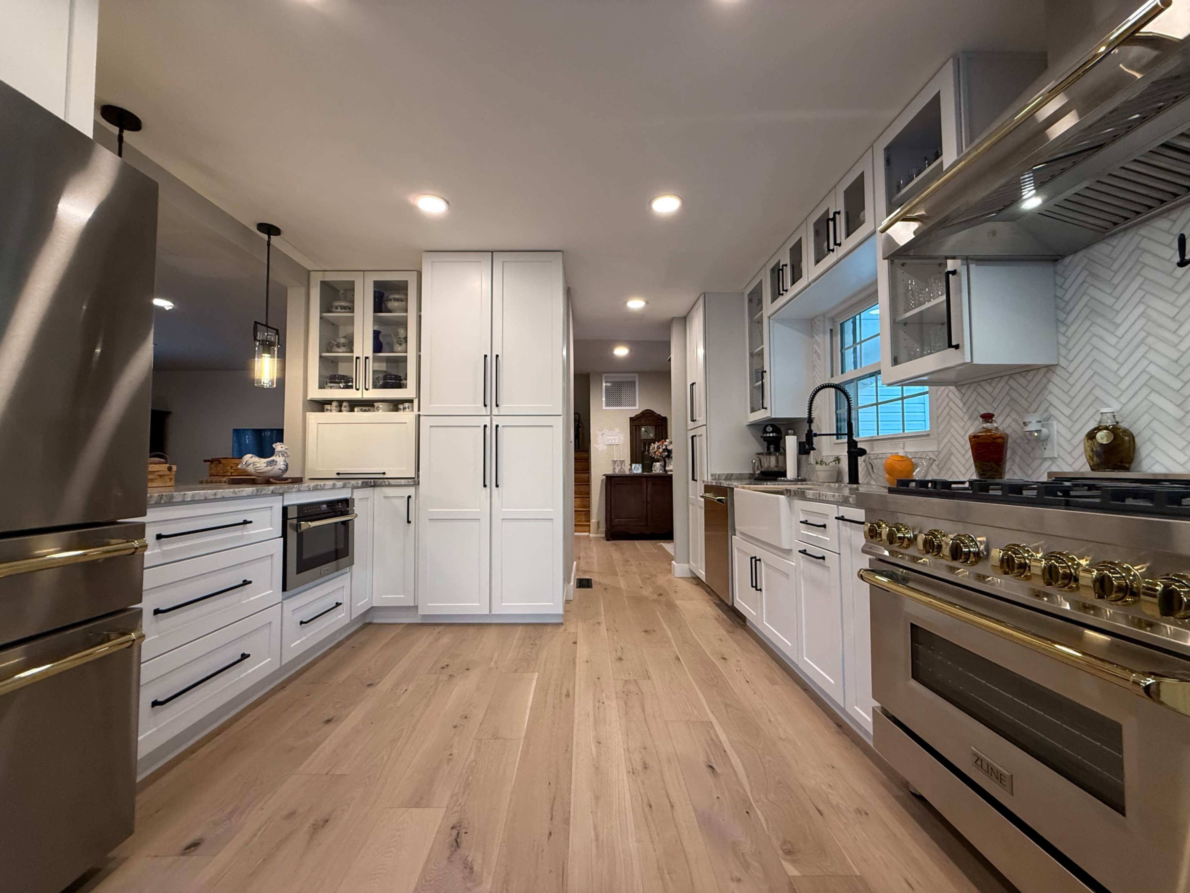 The image shows a modern kitchen featuring white cabinetry, a stainless steel refrigerator, and a gas stove with a hanging hood, all arranged with a wood-like floor.