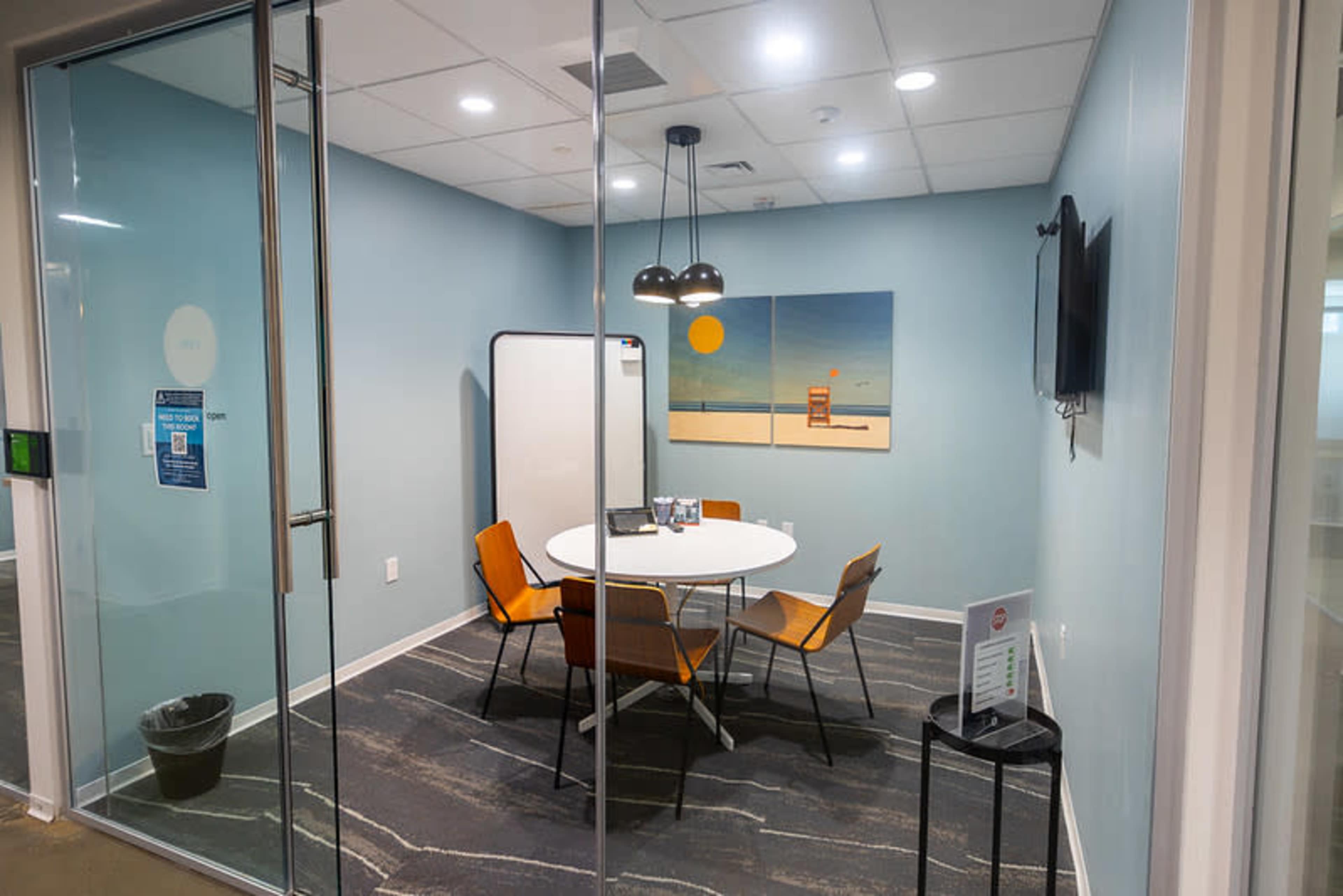 The image shows a glass-walled meeting room with a round white table and four orange chairs, featuring a painting on the wall and a television mounted nearby.