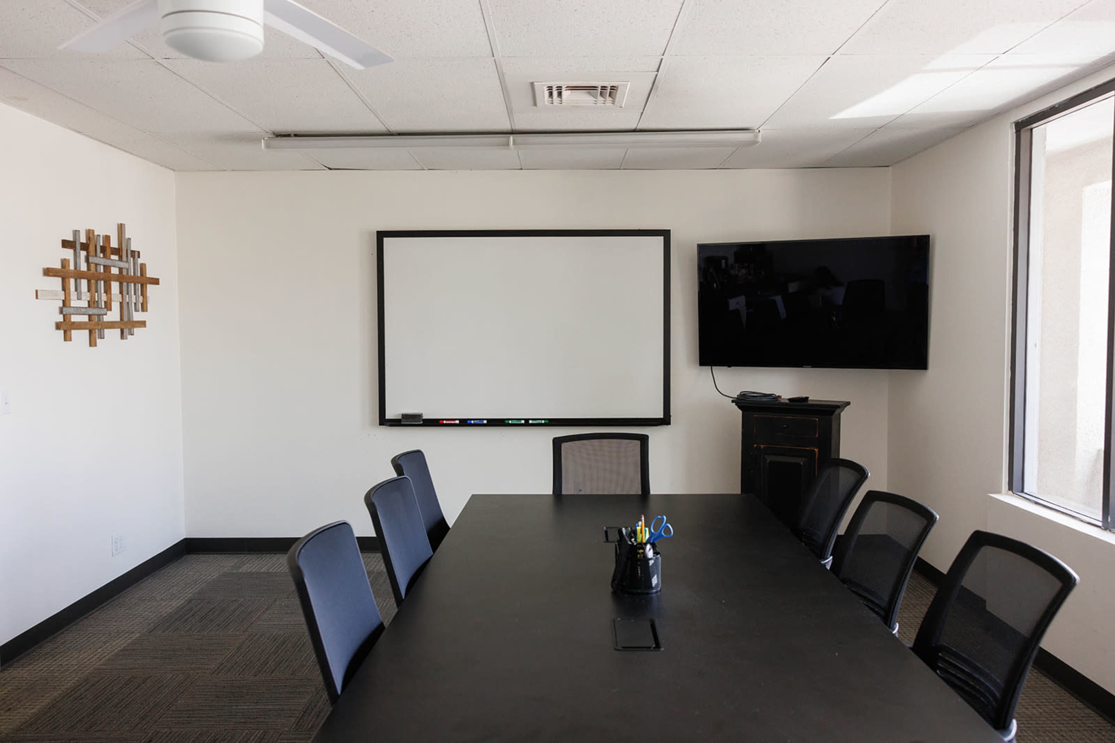 The image shows a minimalist conference room featuring a large black table surrounded by black chairs, a whiteboard, a television screen, and a wall decoration.