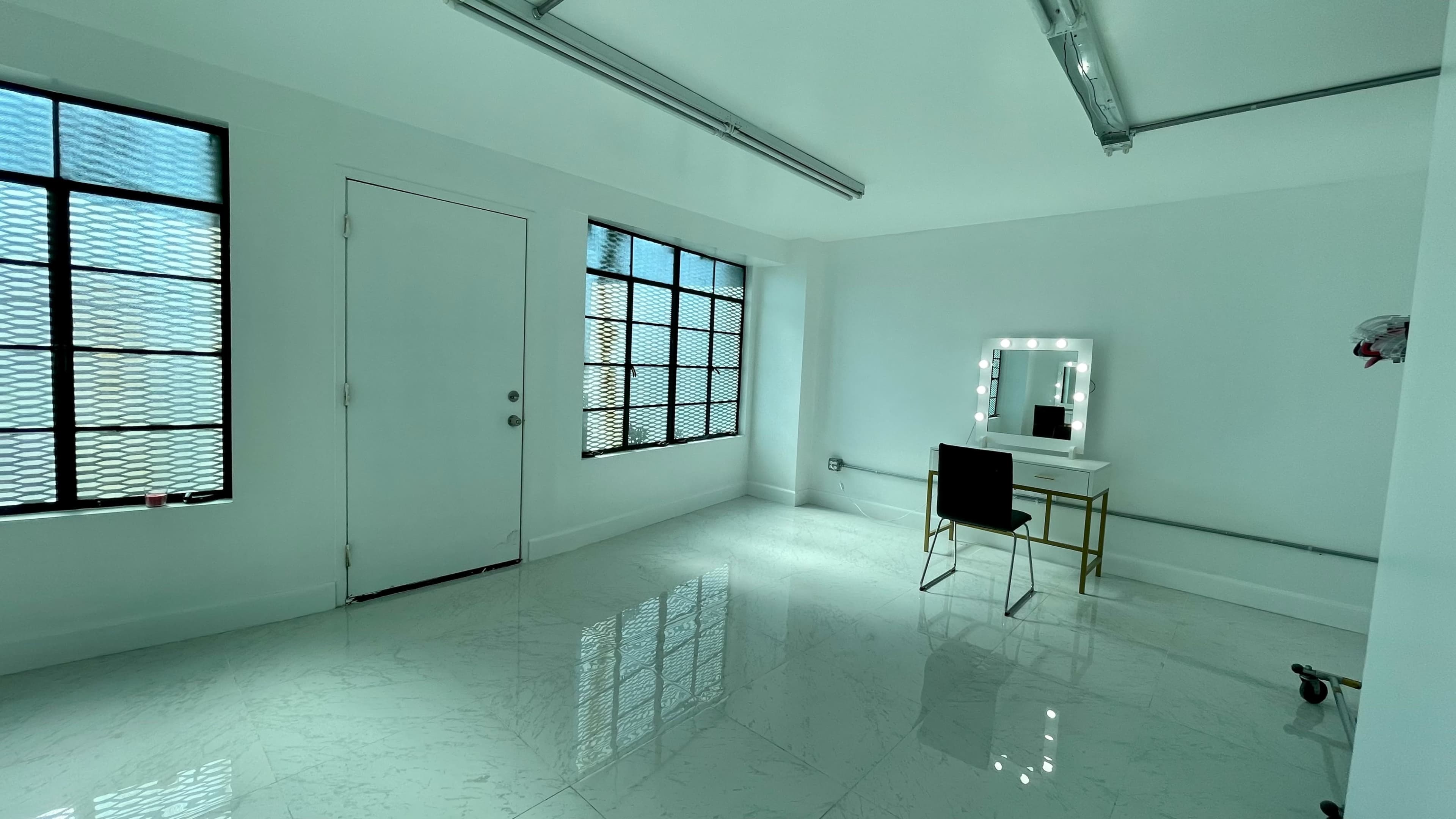 A brightly lit room with white walls, a large mirror on a vanity with lights, and a black chair, featuring two windows with metal grilles.