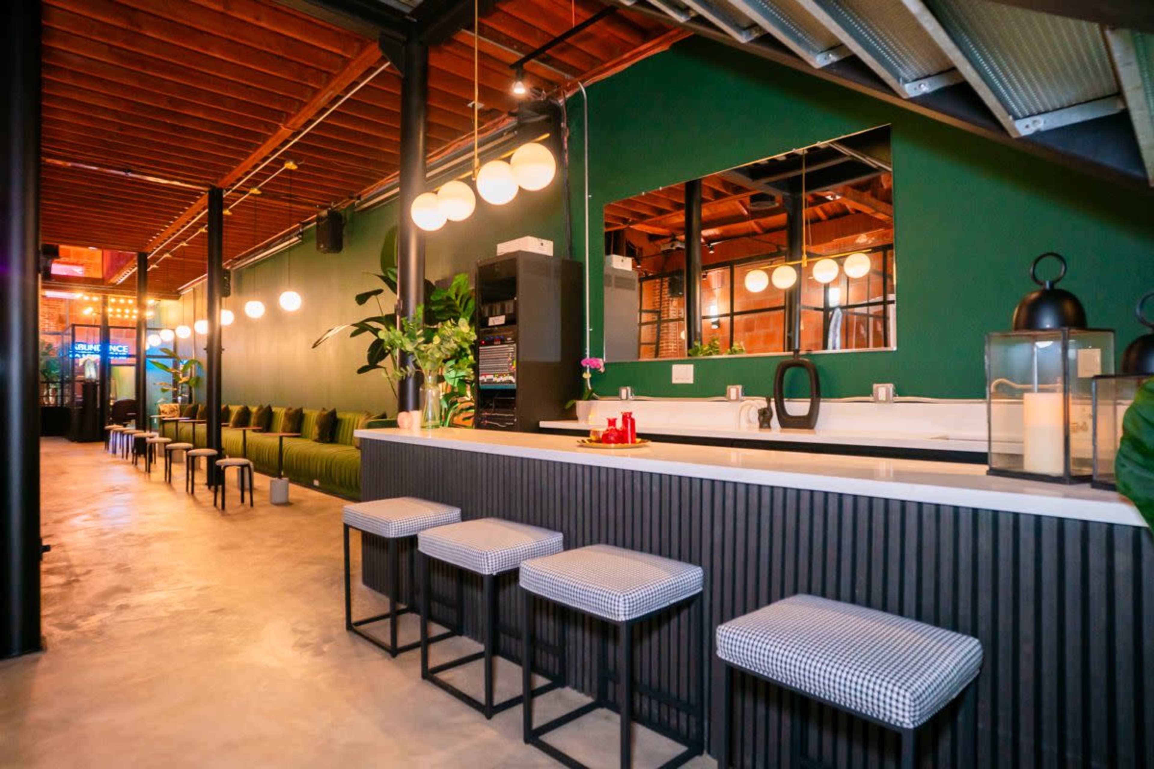 The image shows a modern bar area with a sleek counter, several stools, and green walls illuminated by soft lighting, leading to a seating area in the background.