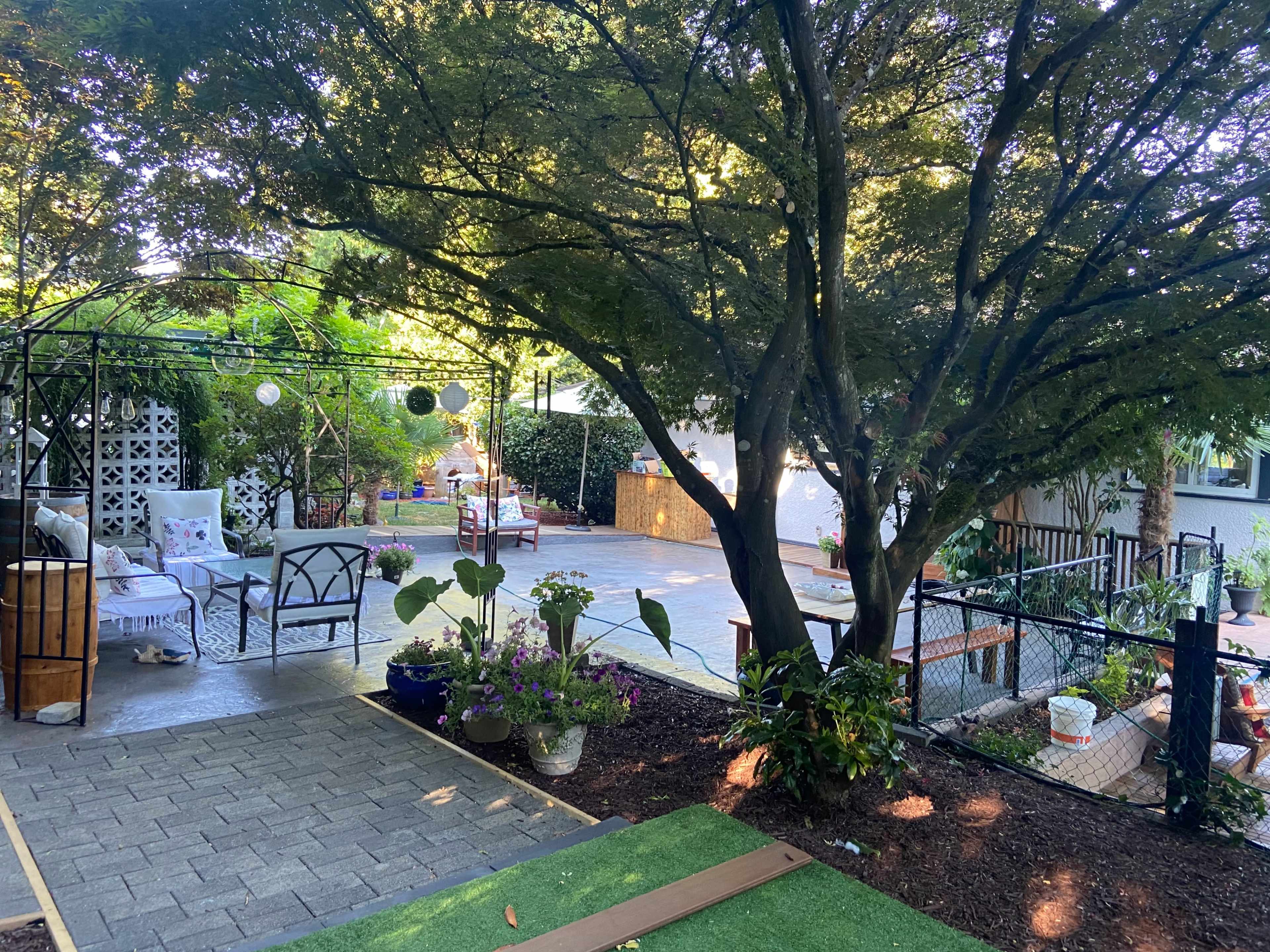 A landscaped garden area with a patio, surrounded by greenery, featuring outdoor seating and a fence.