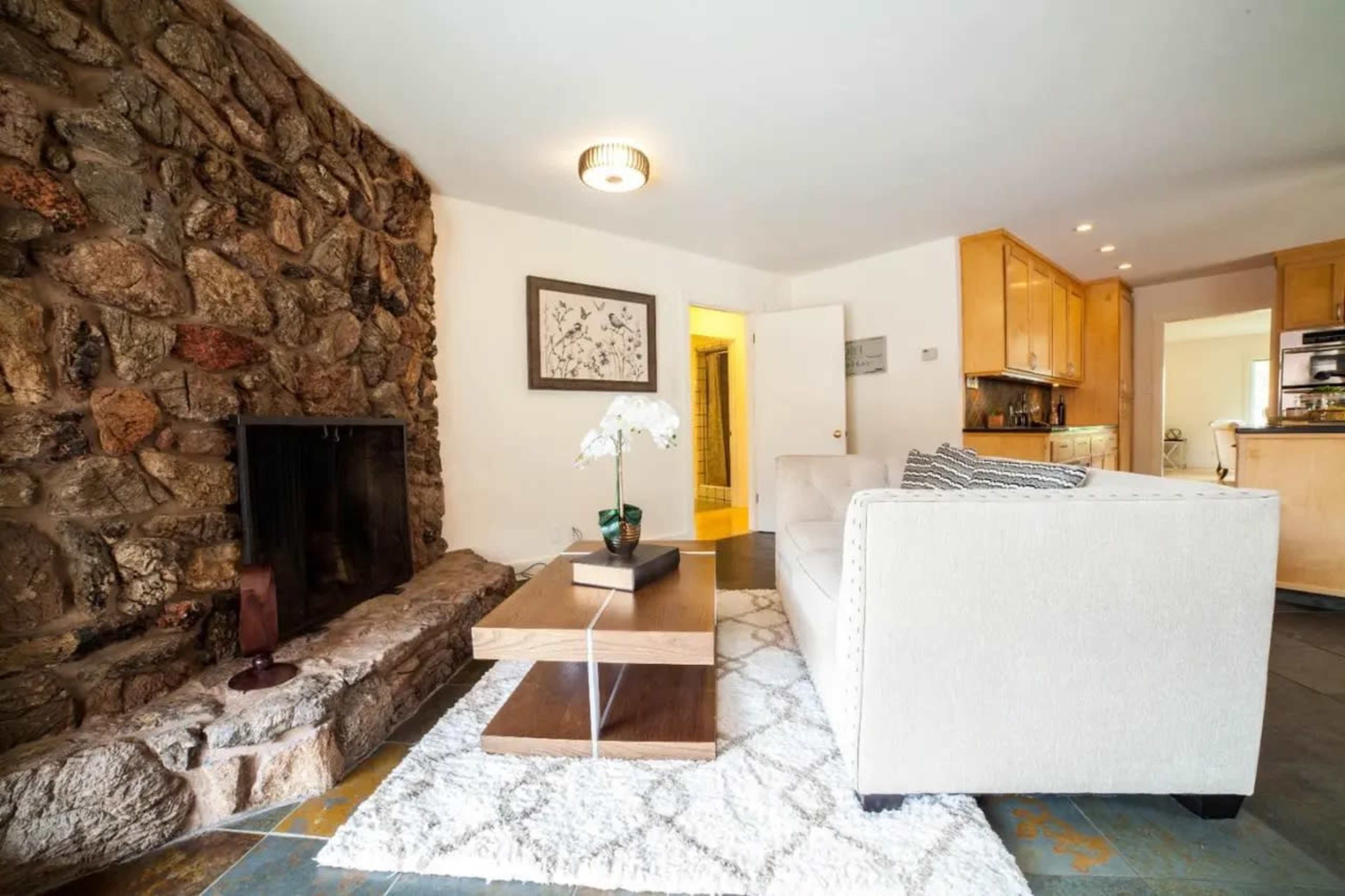 The scene features a living room with a stone accent wall, a beige sofa, a wooden coffee table, and a visible kitchen area through an open doorway.