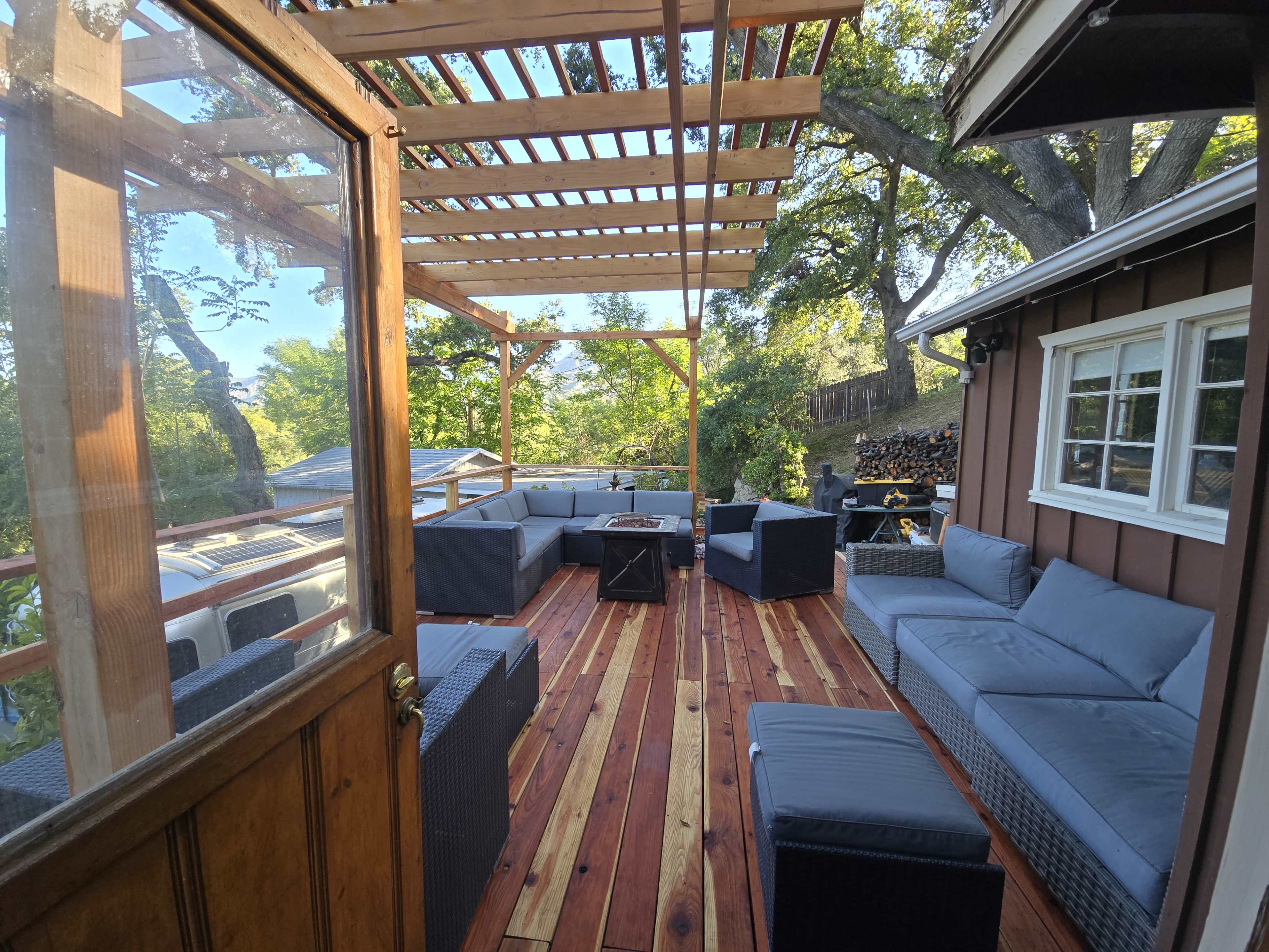 The image shows a wooden deck with modern seating arranged around a central table, enclosed by a wooden frame structure and overlooking a green outdoor landscape.