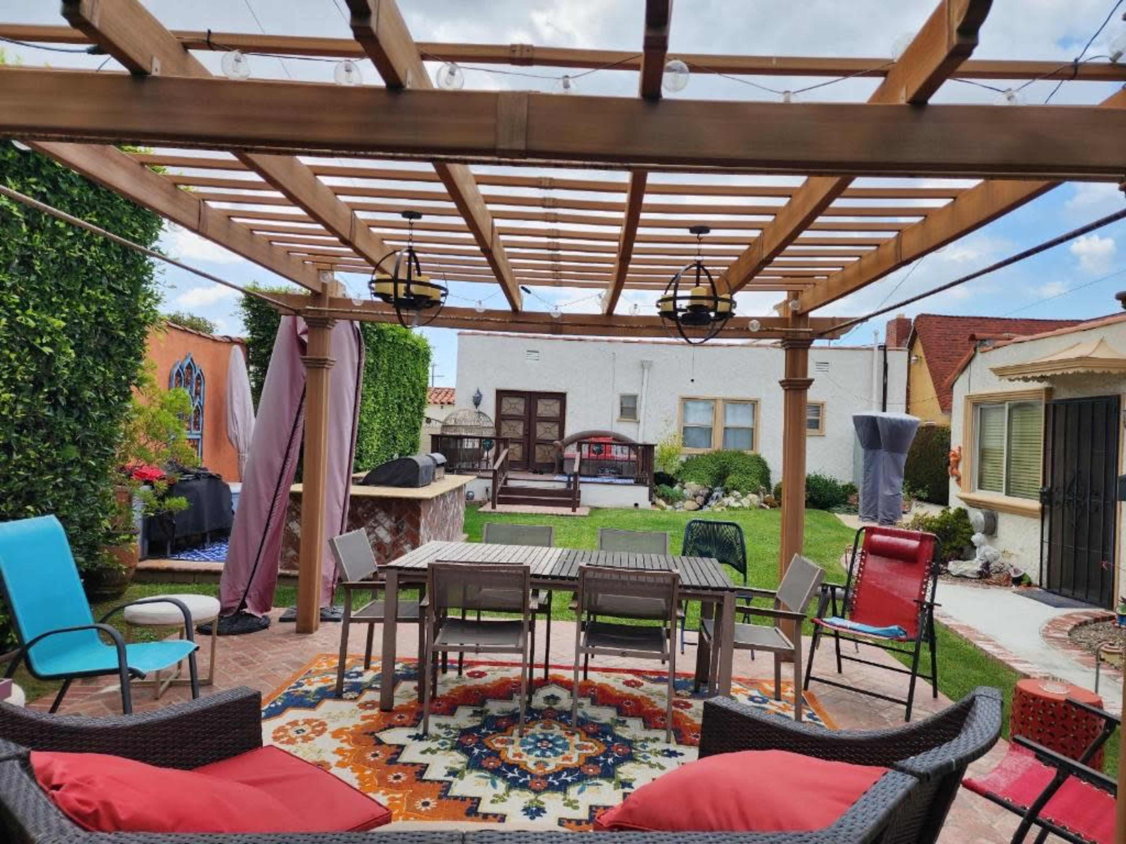 A backyard patio features a wooden pergola with seating, a dining table, and a colorful area rug, surrounded by lush greenery and outdoor amenities.