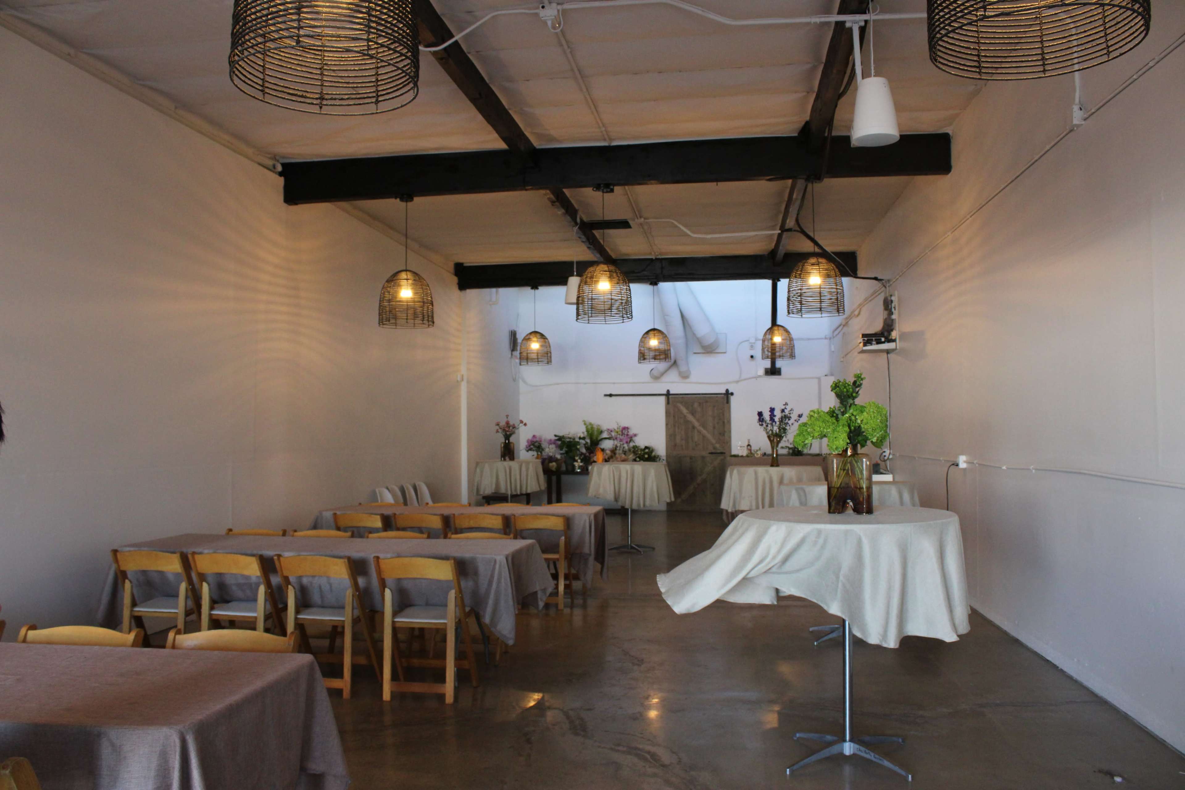 The image shows a spacious, well-lit event room with wooden tables covered in light-colored tablecloths and decorative plants arranged on them.