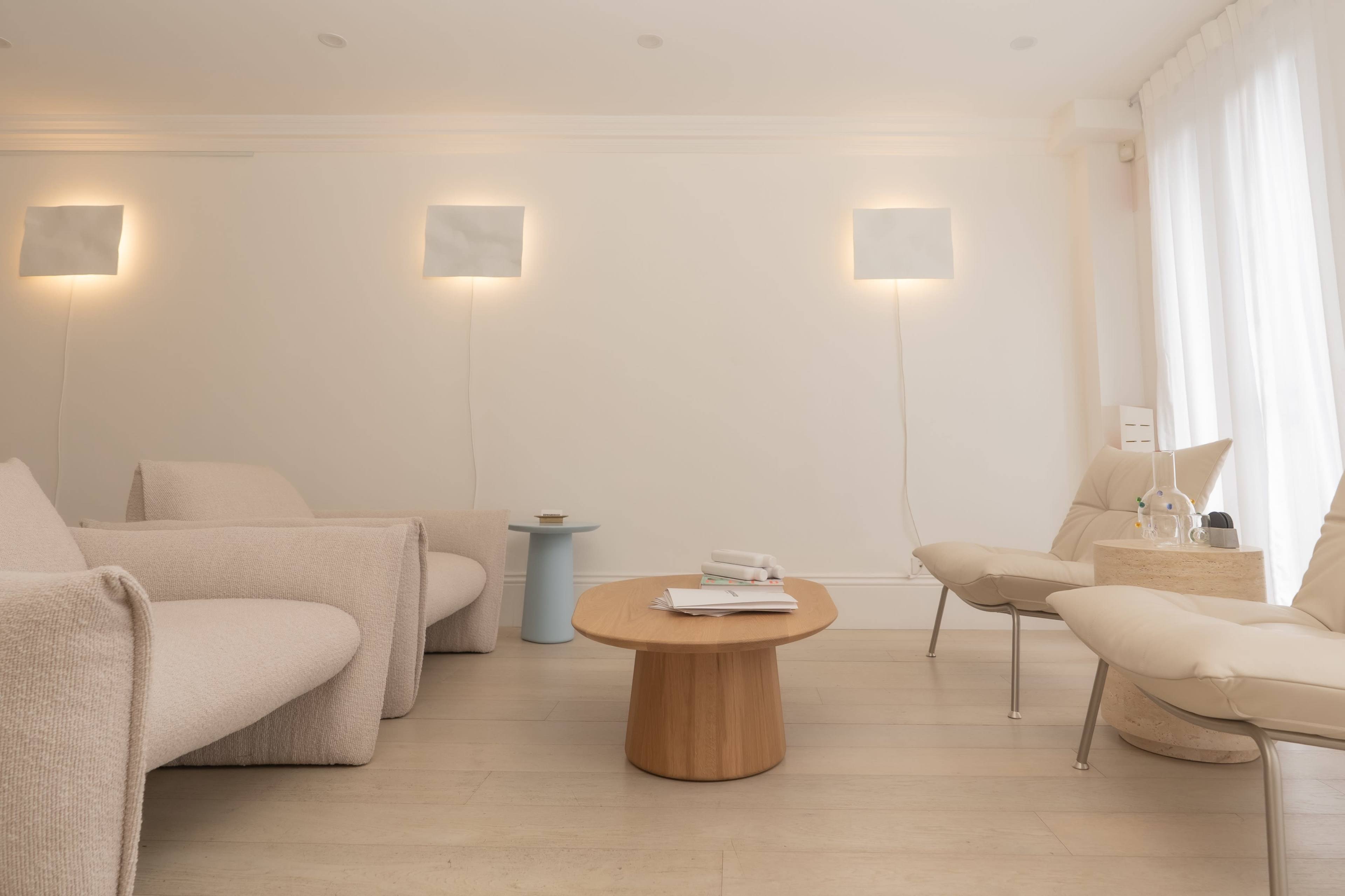 The image shows a minimalistic interior of a living room with two light-colored armchairs, a circular wooden coffee table, and soft lighting on the walls.