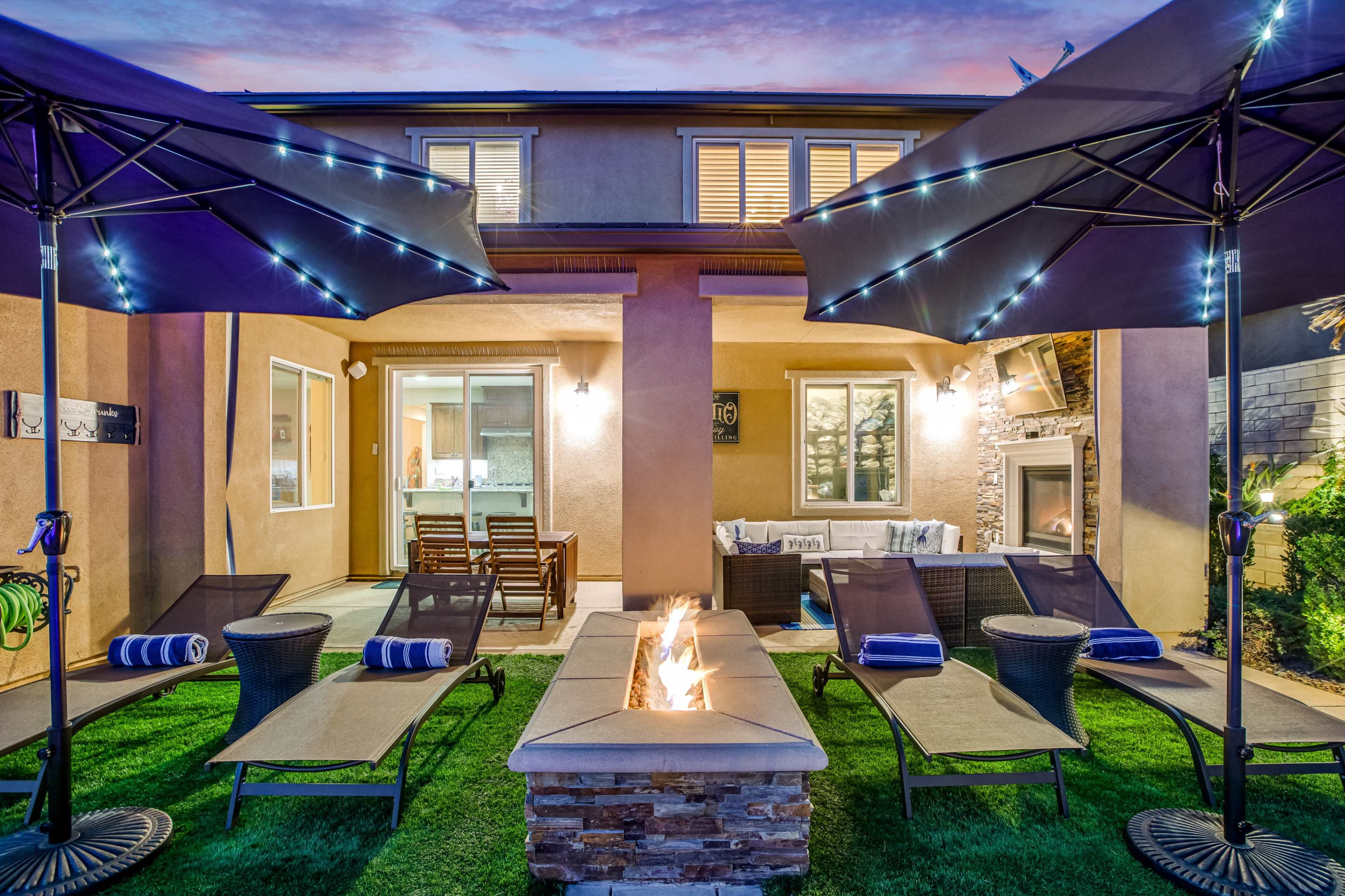 The image shows a backyard patio with lounge chairs, a fire pit, and illuminated umbrellas, overlooking a modern home.