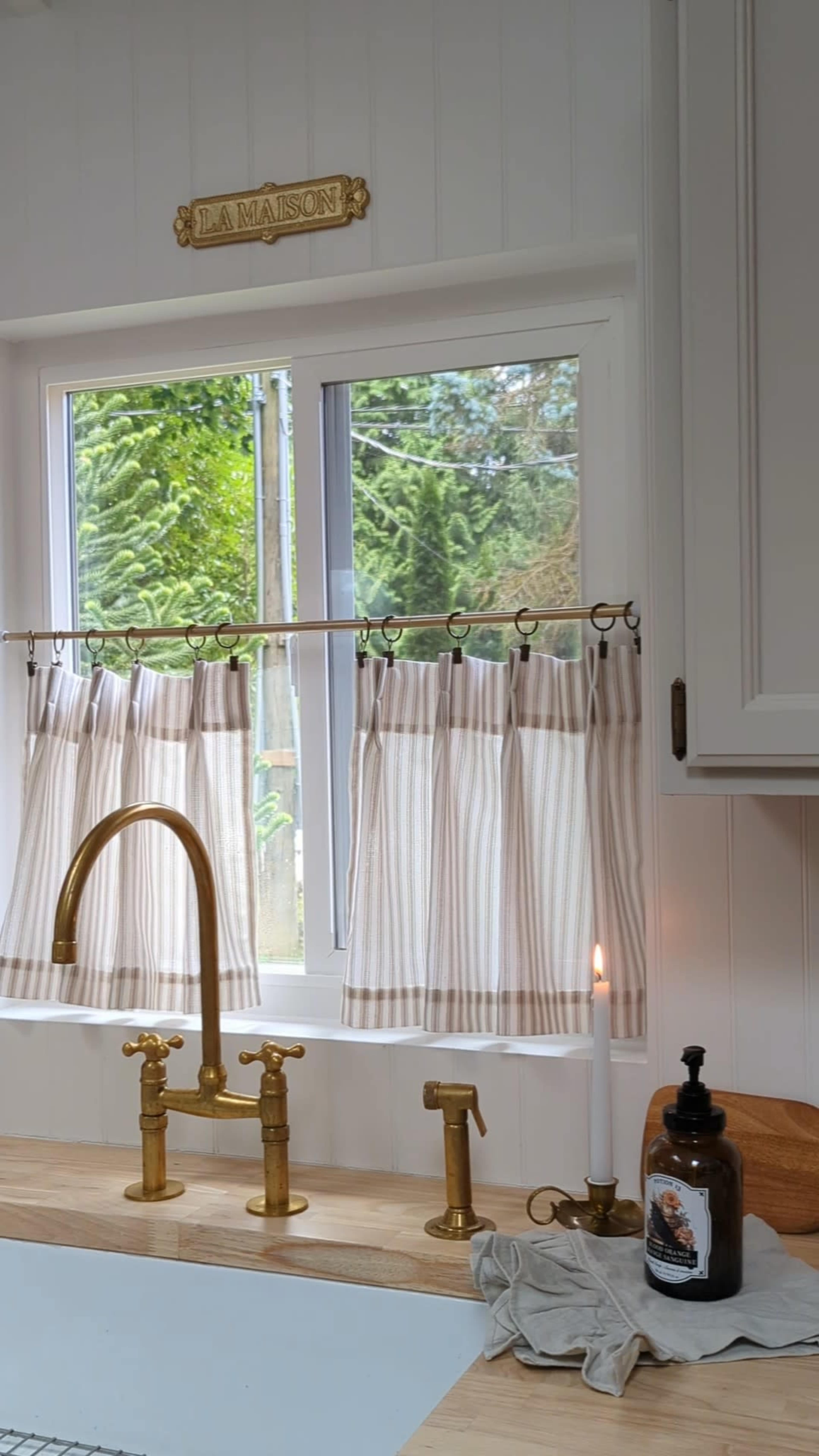 The image shows a kitchen window adorned with striped curtains, a brass faucet, and a candle on the countertop.