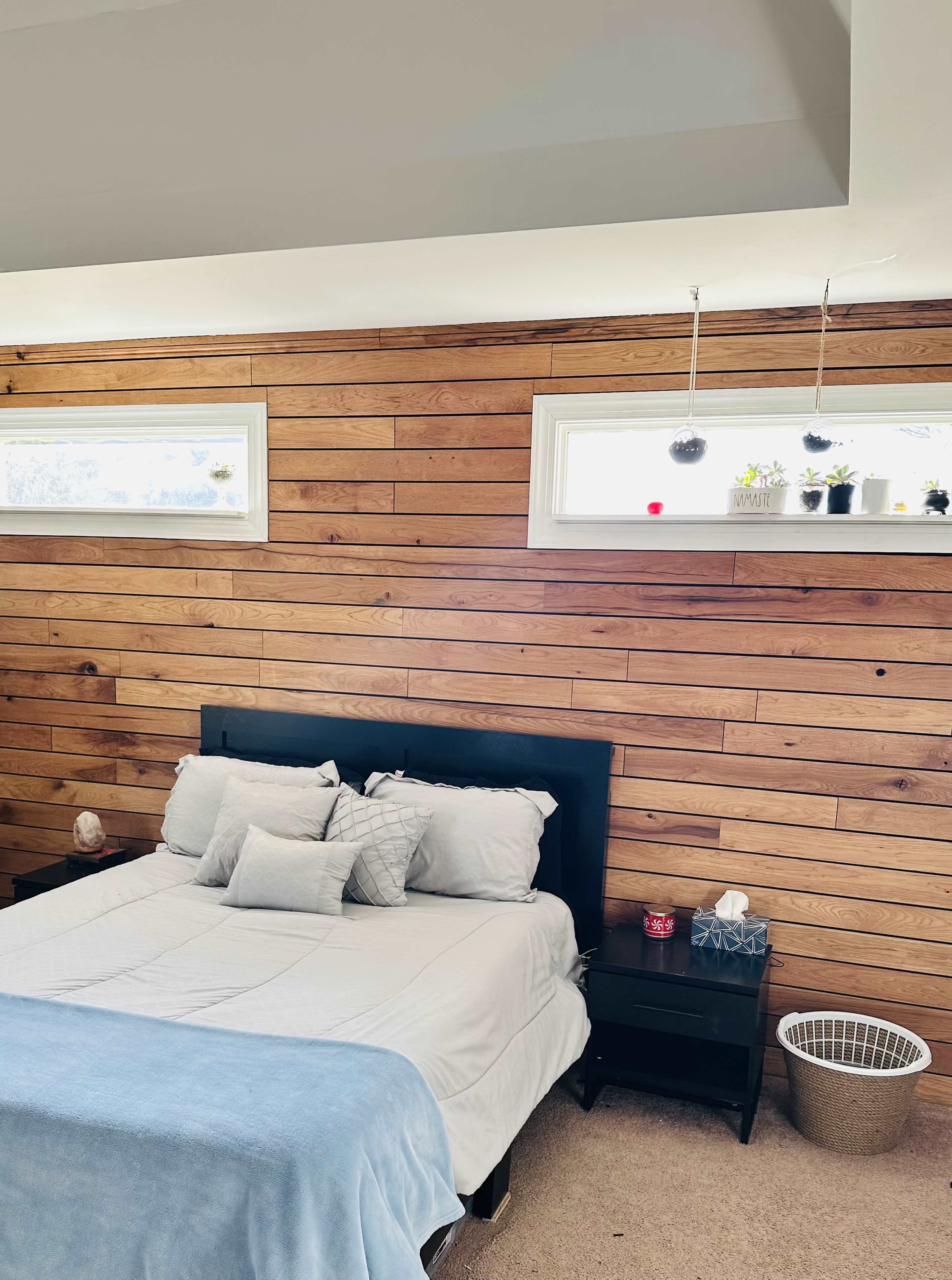 A neatly made bed with gray pillows and a light blue throw blanket is positioned against a wood-paneled wall, accompanied by two window sills adorned with small plants.