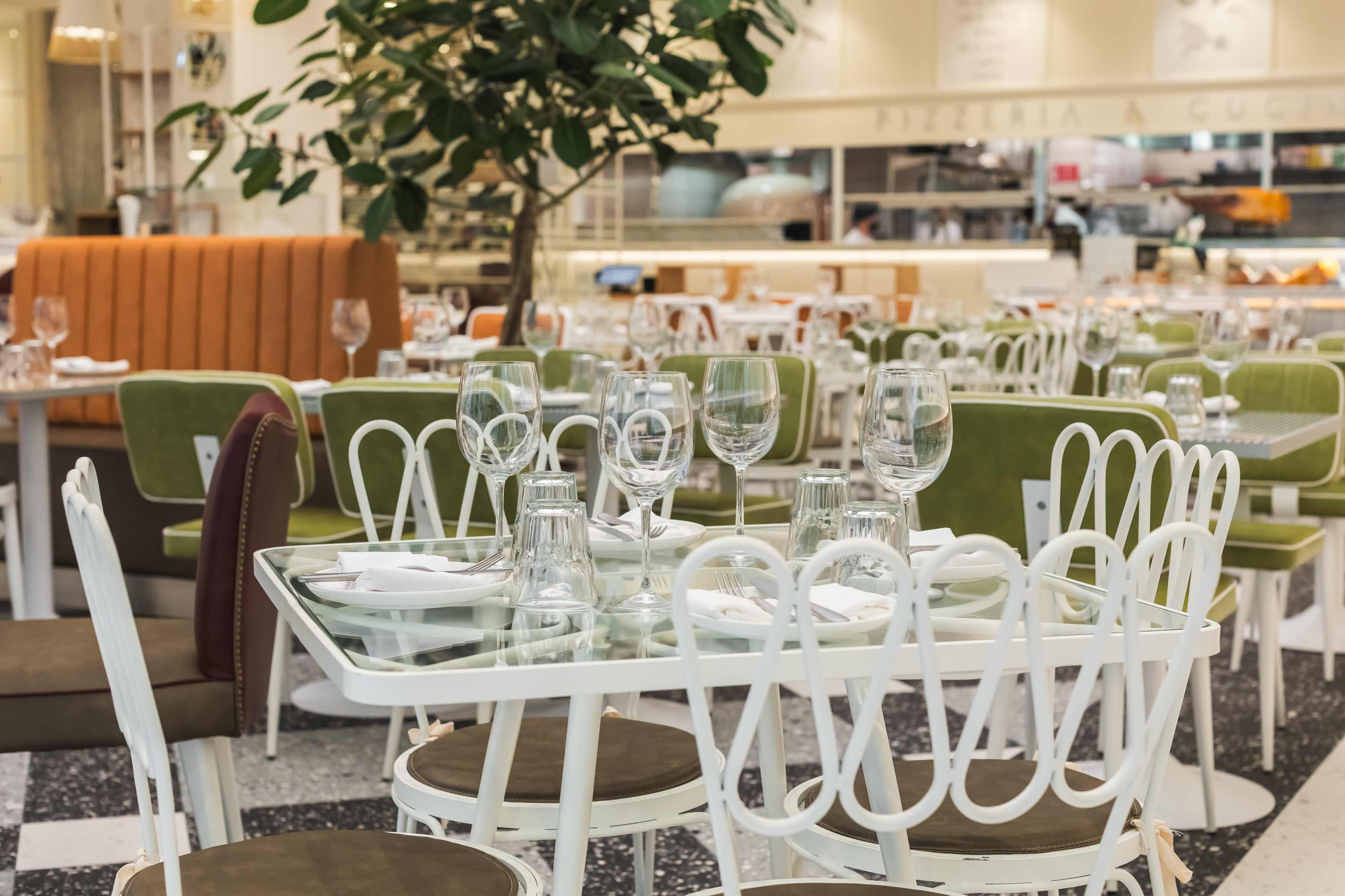 The image shows a modern restaurant interior with neatly set tables, a vibrant green and orange color scheme, and a large indoor tree.