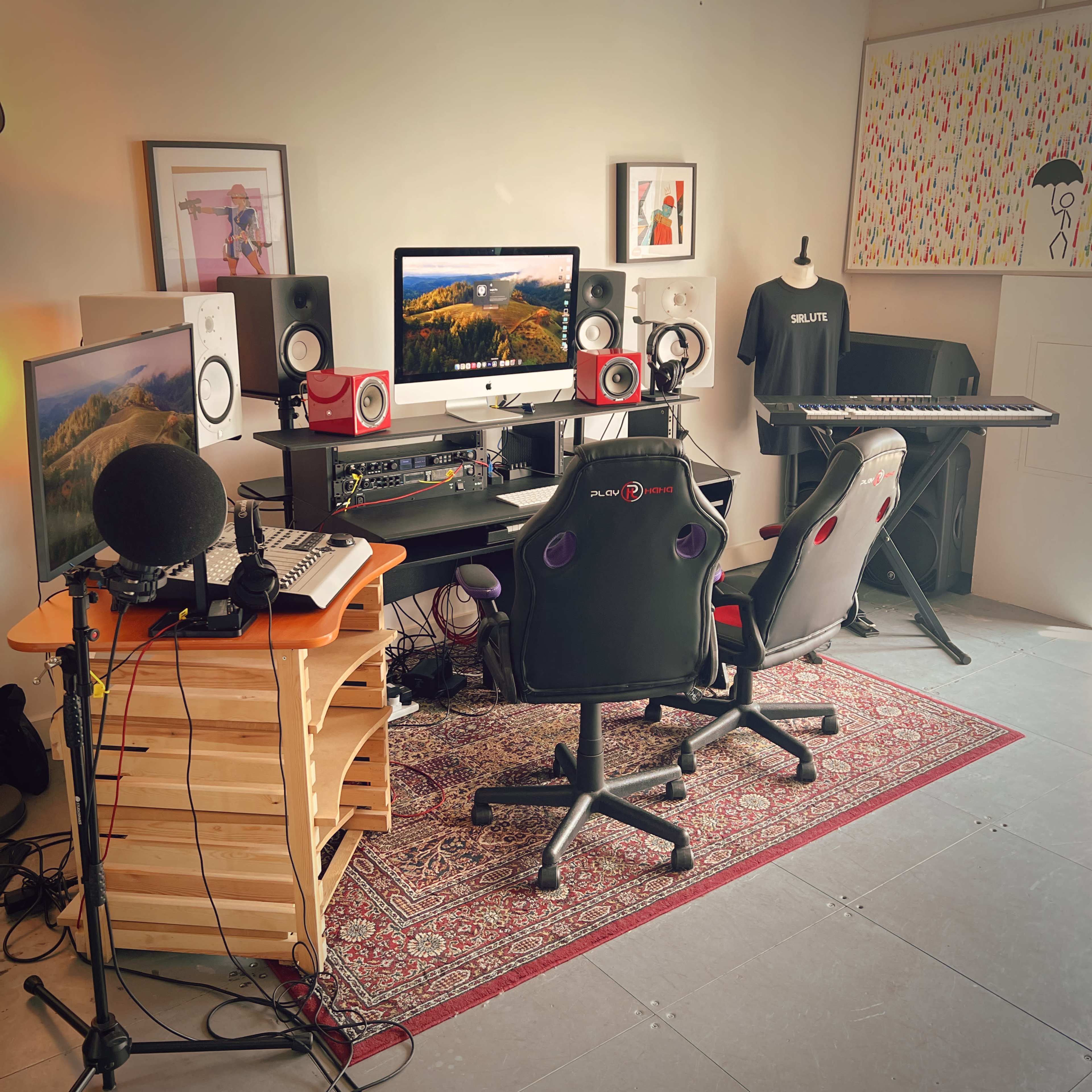 A music production studio features a computer setup with large monitors, speakers, and a keyboard, surrounded by chairs and equipment on a patterned rug.