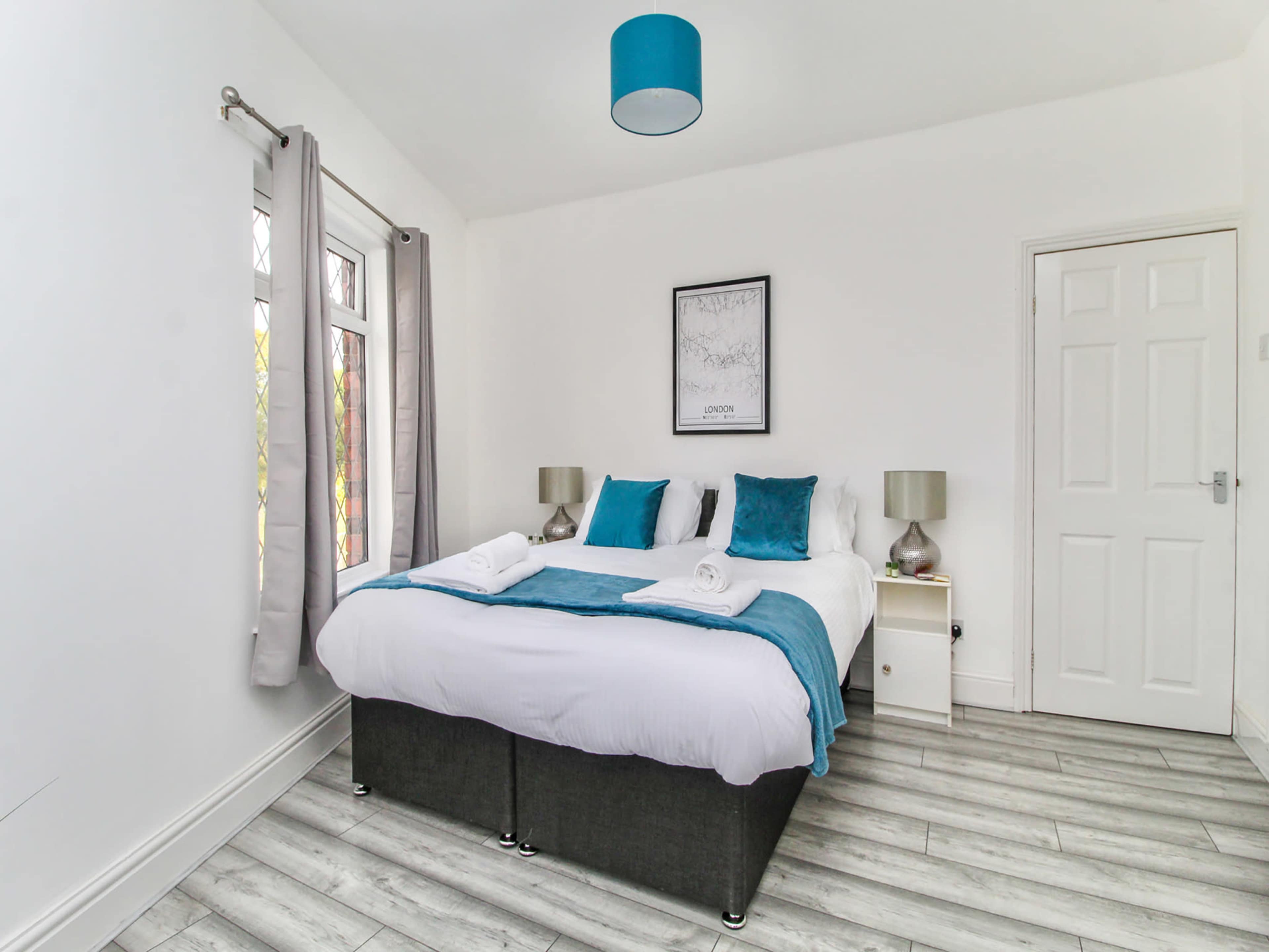The image shows a well-lit bedroom featuring a double bed with white linens, teal accent pillows, and a matching throw, along with bedside tables and a window with gray curtains.