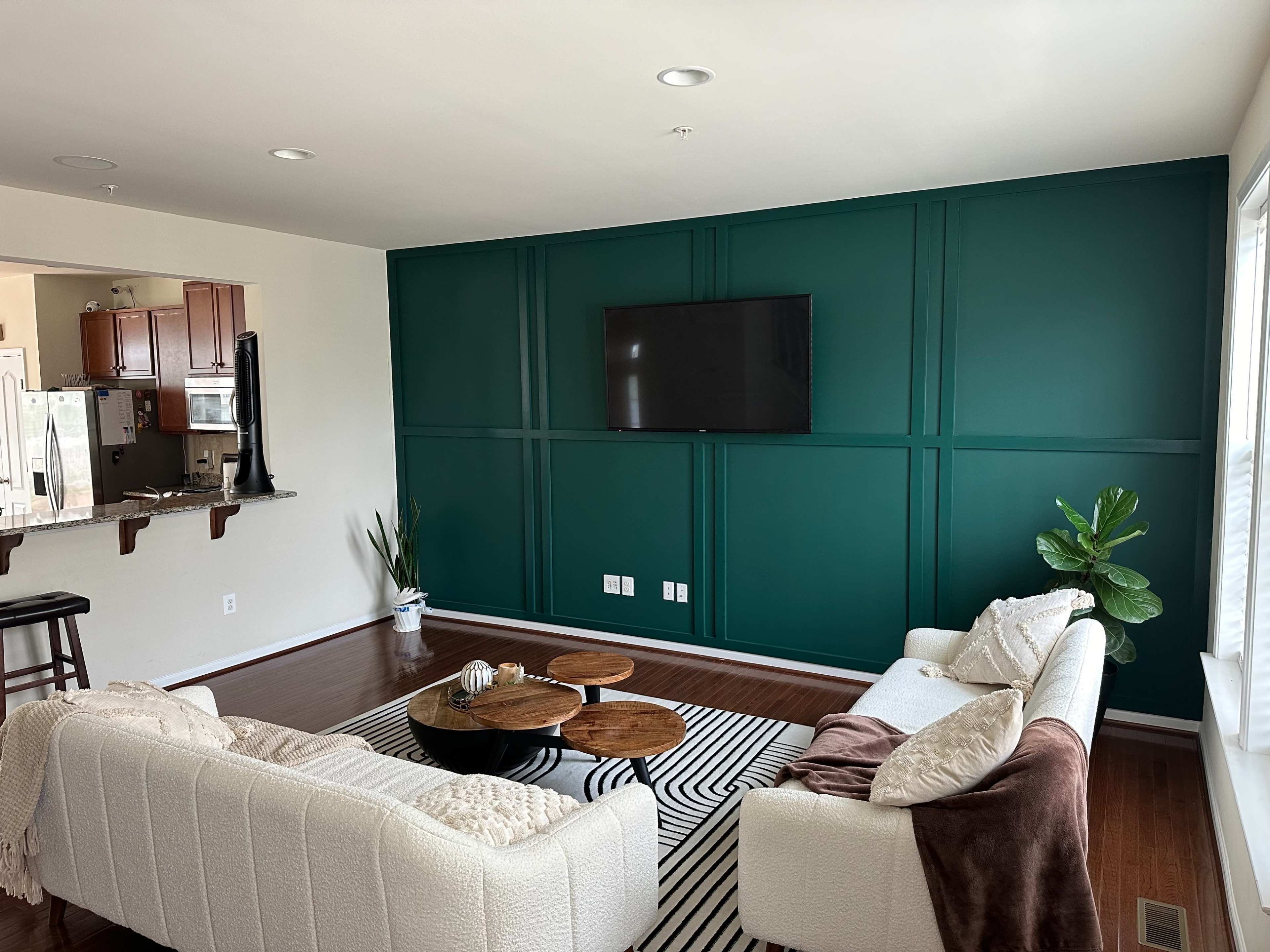 A living room with a green accent wall, a flat-screen TV mounted on it, a white sectional sofa, a striped rug, and a small plant in the corner.