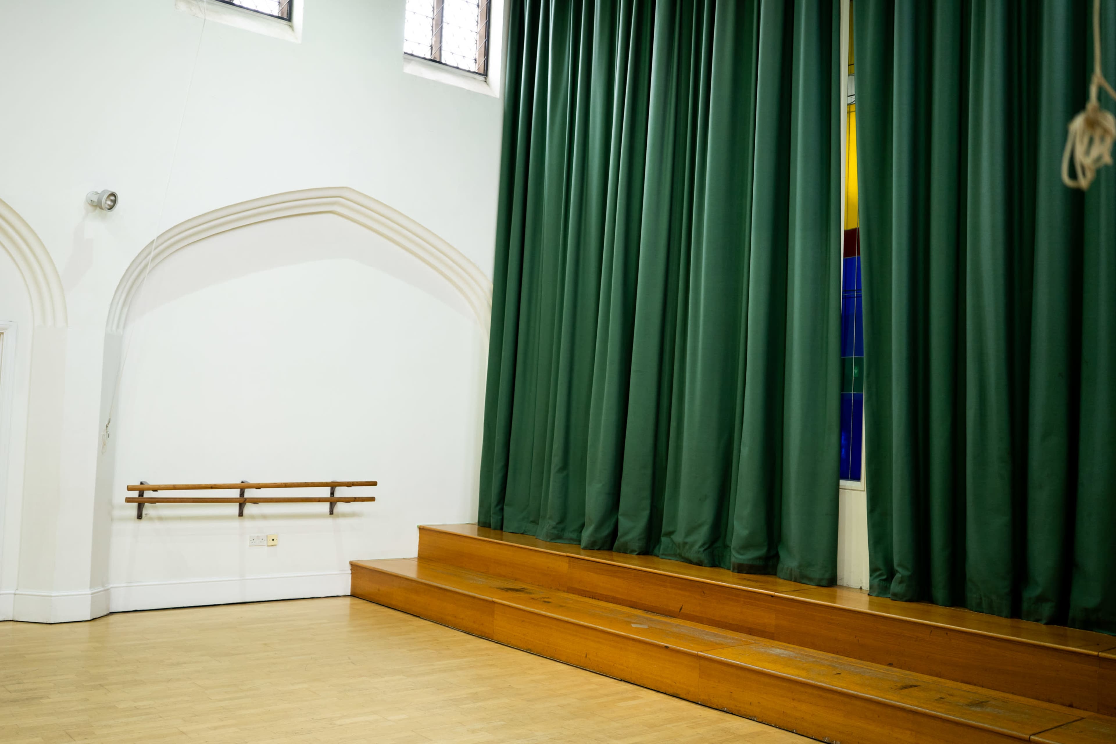 The image shows a corner of a room with wooden steps leading up to a stage, flanked by green curtains and a stained glass window.