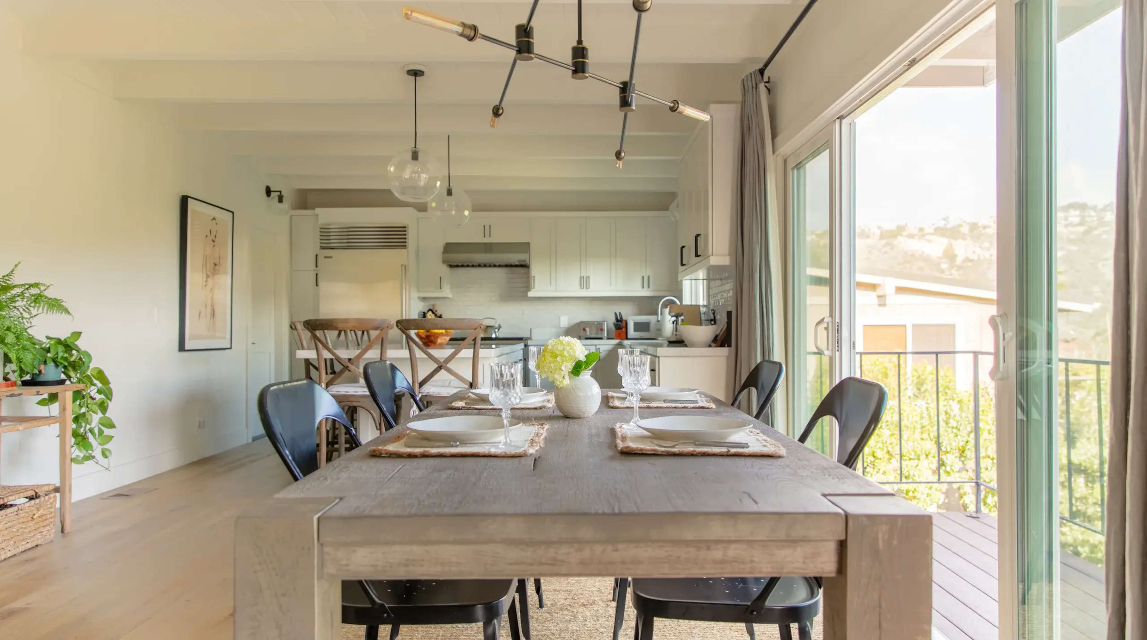 A wooden dining table with seating for six is set in a bright room that connects to a modern kitchen through large windows.
