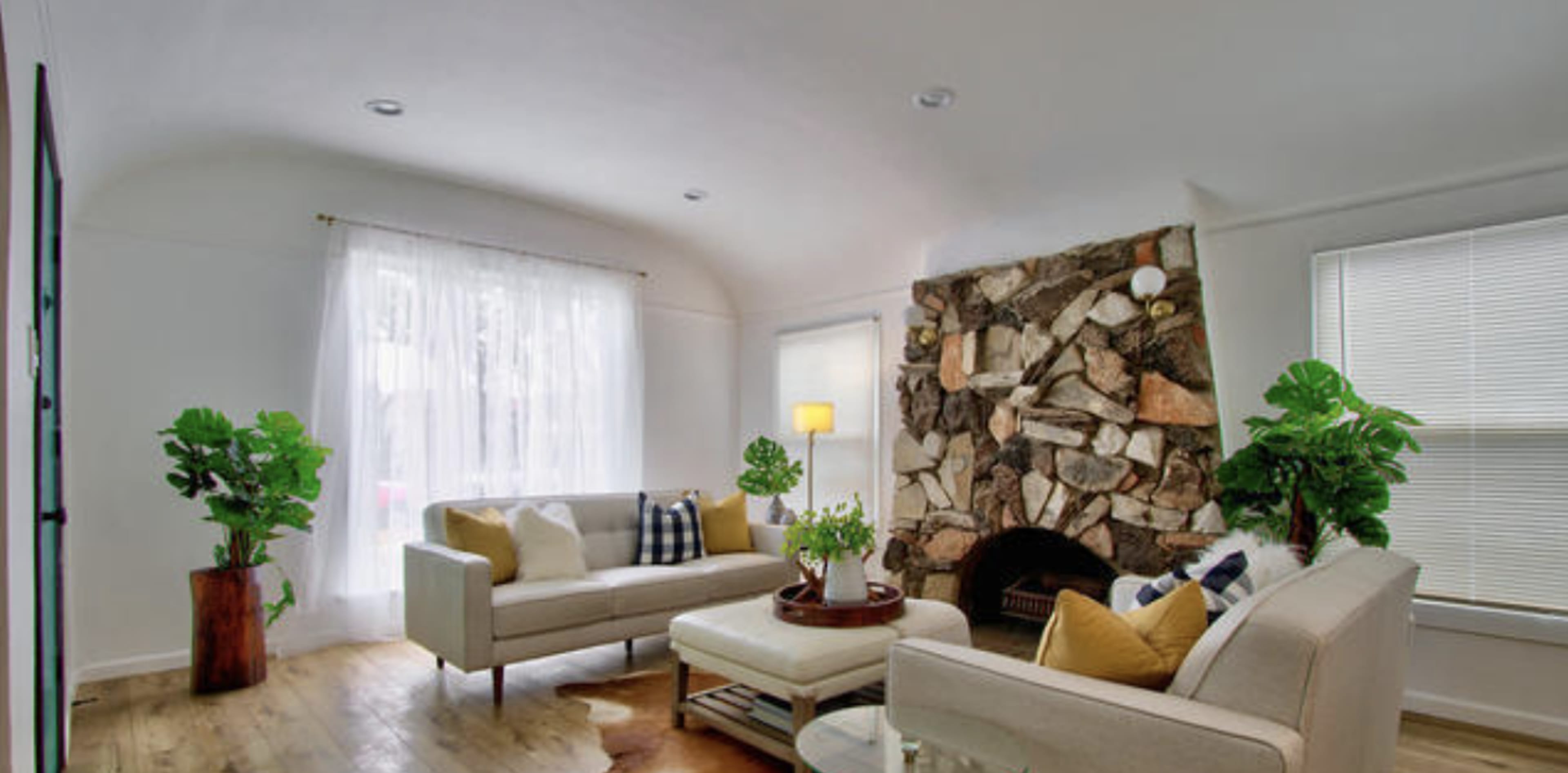 The living room features a stone fireplace, two sofas with decorative pillows, a coffee table, and plants adding greenery to the space.