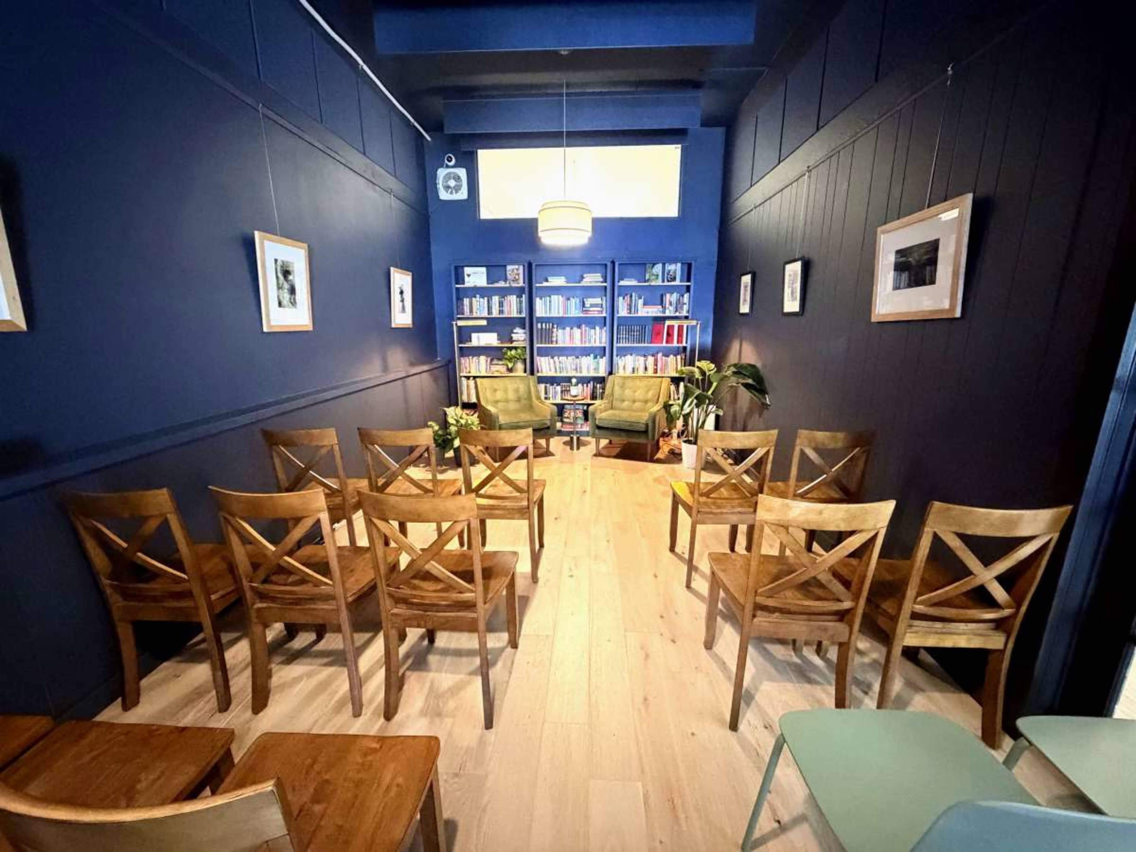 The image depicts a small, dimly lit room with dark blue walls, featuring several wooden chairs arranged in rows facing a pair of green armchairs and a bookshelf filled with books.