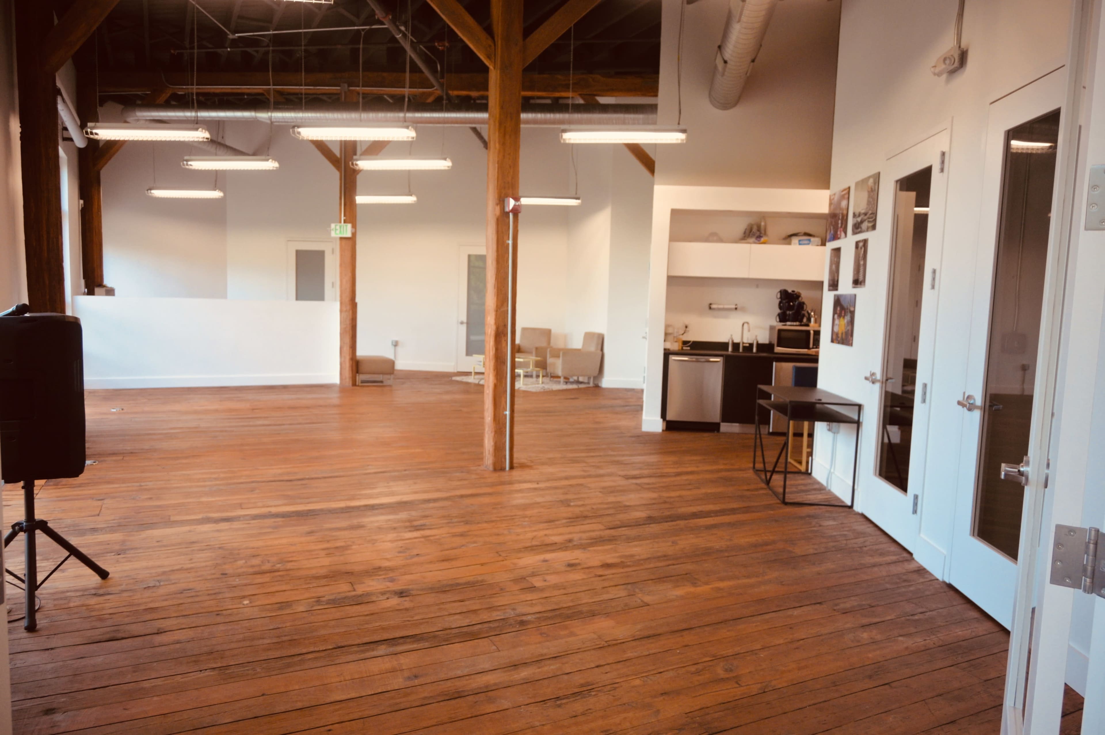 The image shows a spacious, empty room with wooden floors, large windows, and a kitchenette in the corner.