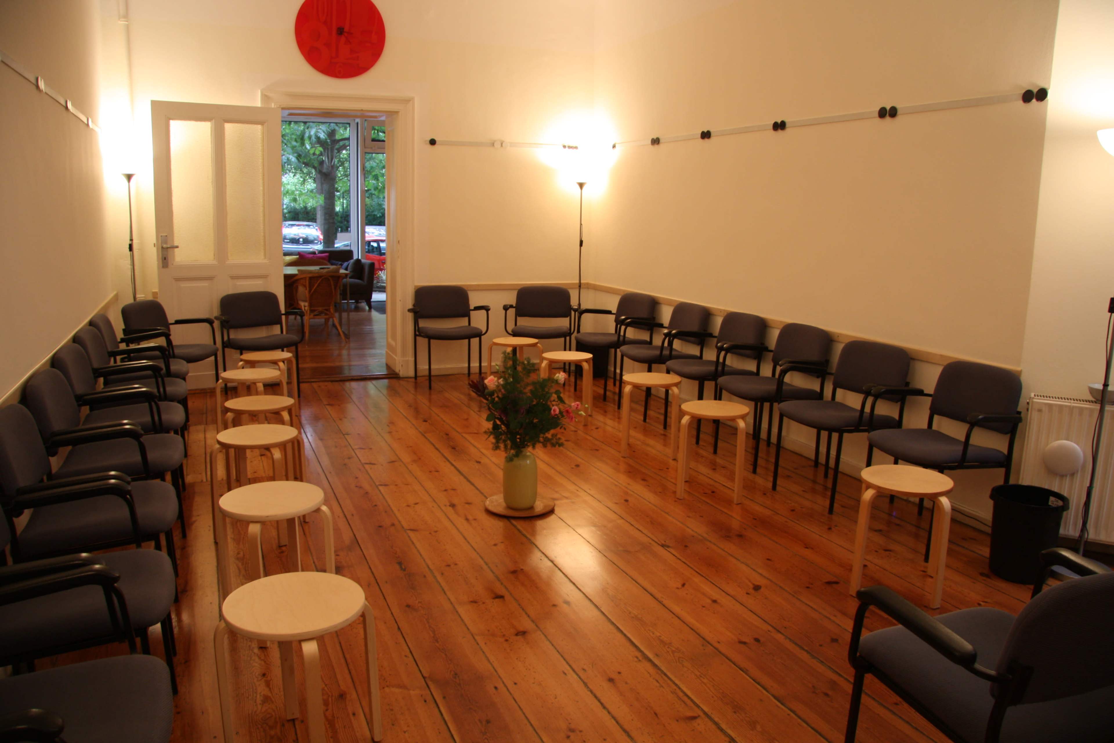 The image shows a spacious room arranged with circular seating, featuring rows of chairs and small wooden tables on a wooden floor.