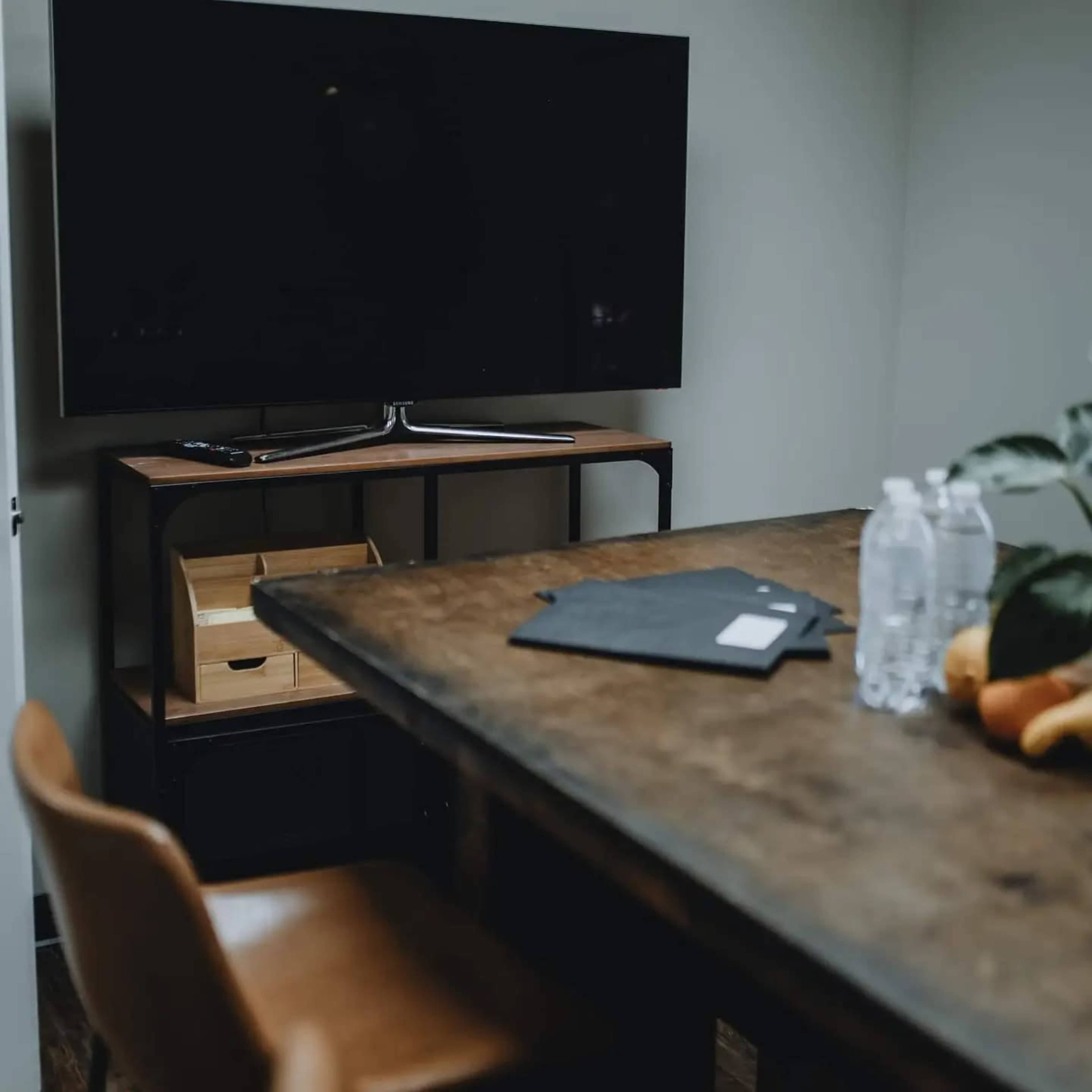 A wooden table is set in a room with a dark television mounted on the wall and a shelf below holding water bottles and a plant.