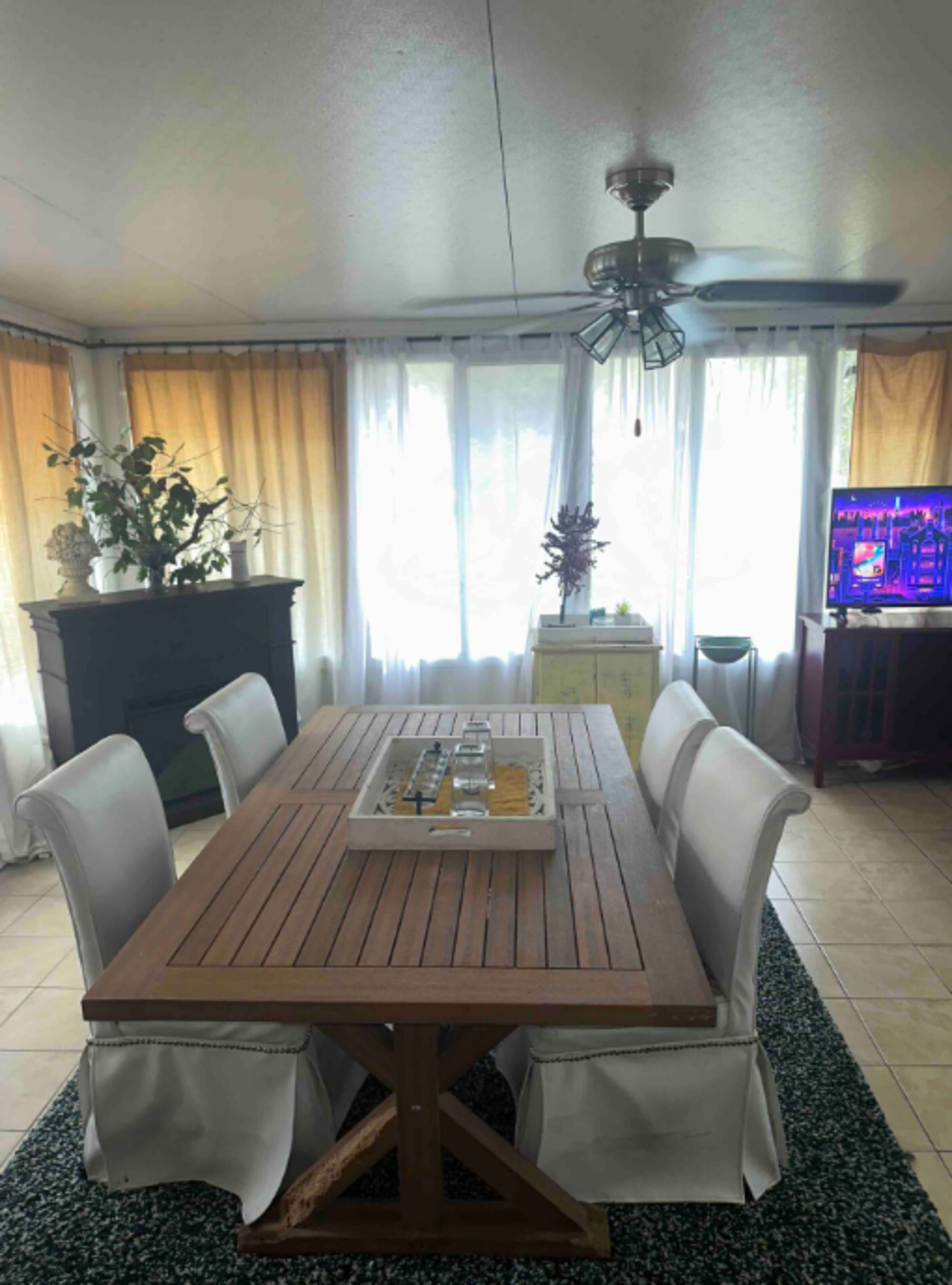 A dining area with a wooden table surrounded by white upholstered chairs, large windows with sheer curtains, and a TV in the corner.