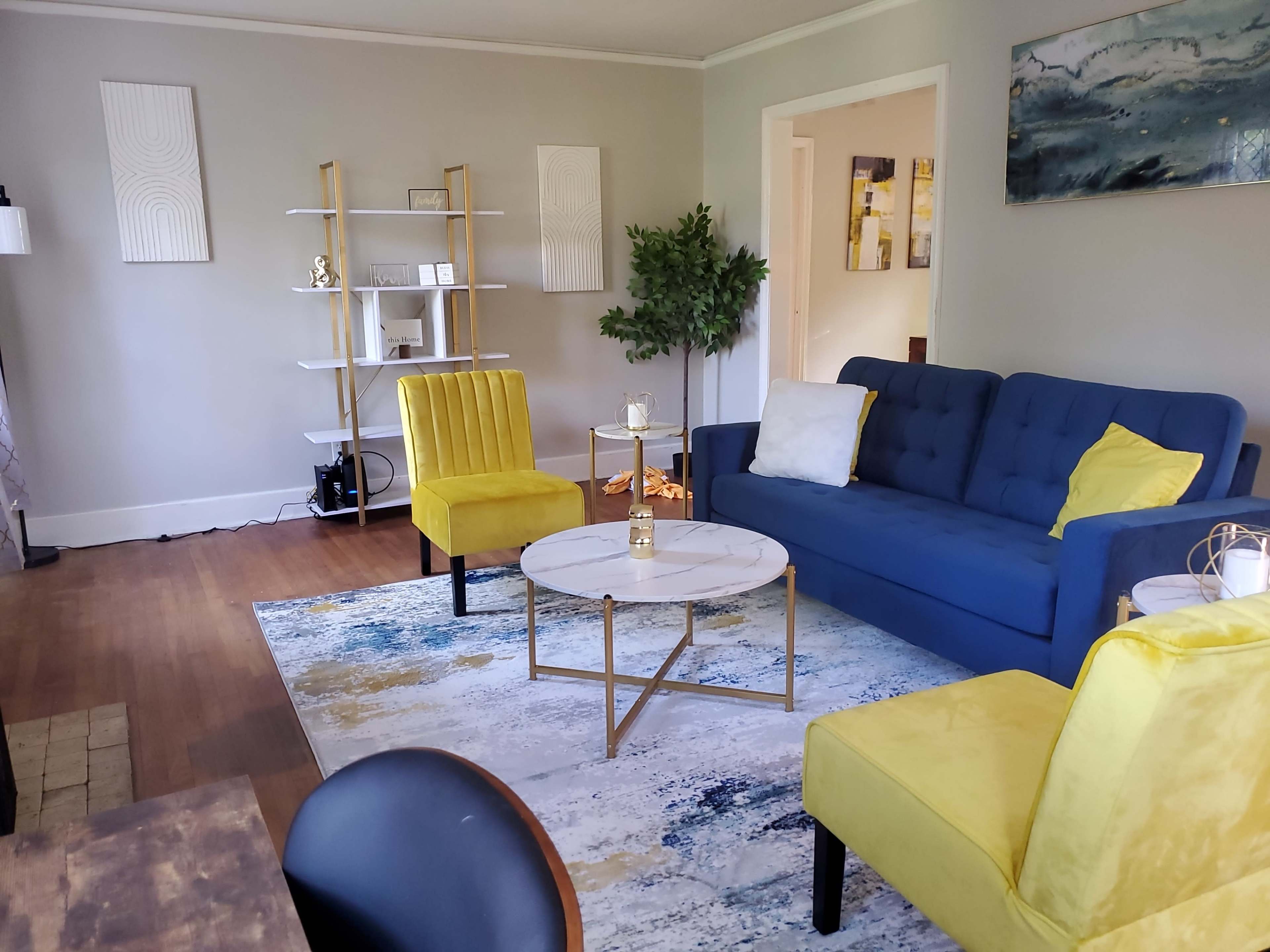 The living room features a blue sofa, two yellow chairs, a round coffee table, and a decorative rug, all arranged around a bookshelf and a potted plant.