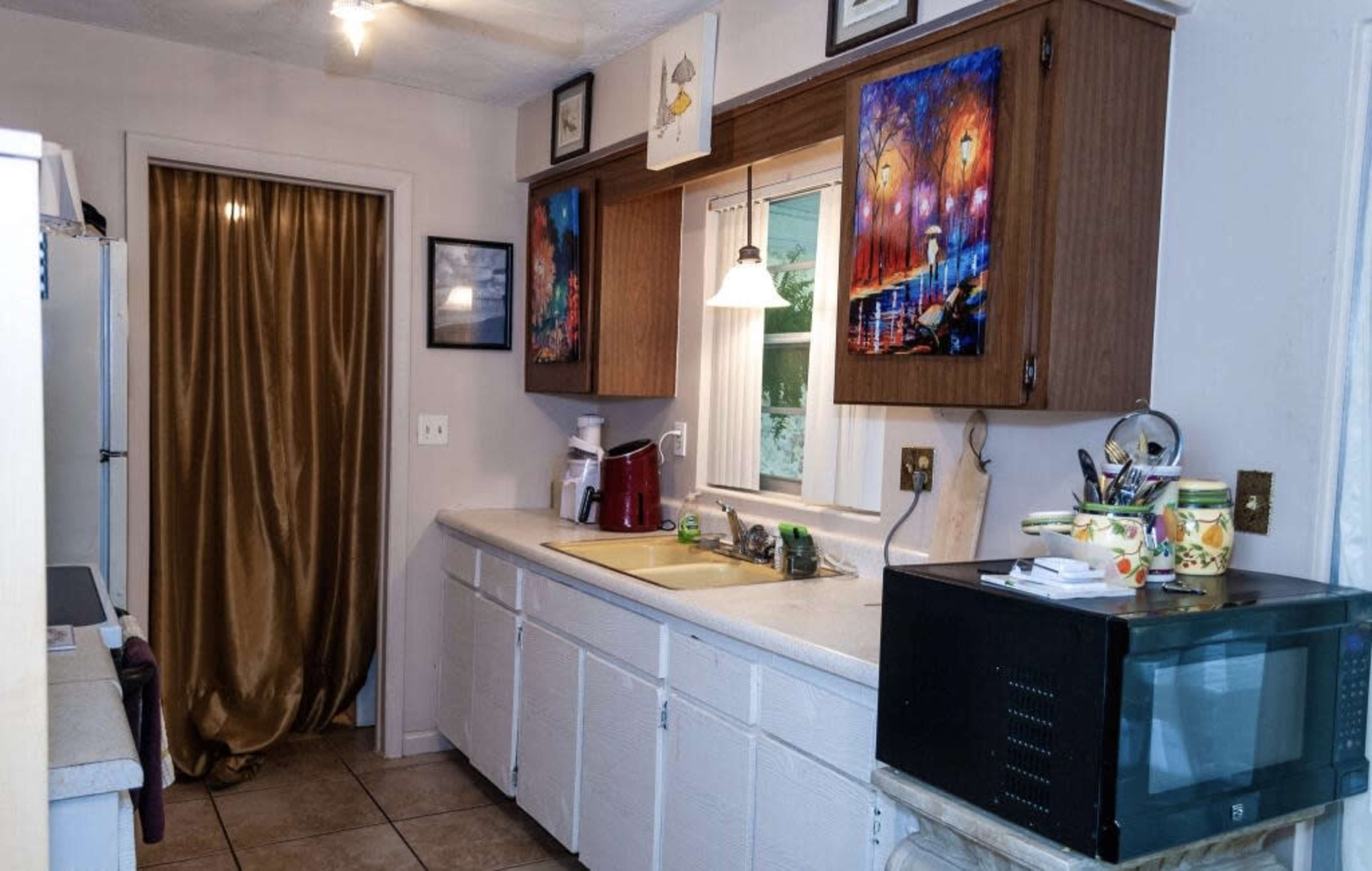 A kitchen features a countertop with a sink, a microwave, and artwork on the cabinets, while a curtain covers a doorway at the far end.